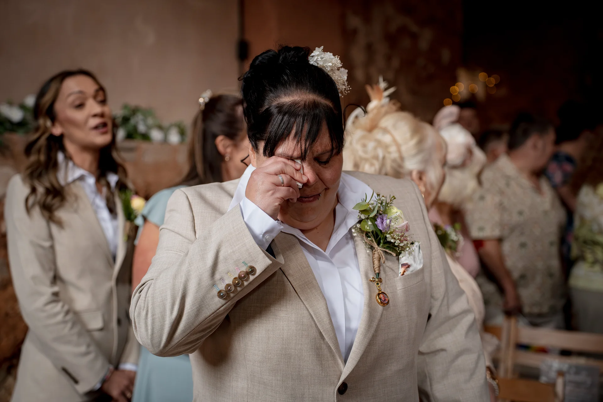 An emotional guest in a beige suit wipes away tears during a wedding ceremony, wearing a floral buttonhole with a small framed photo attached.