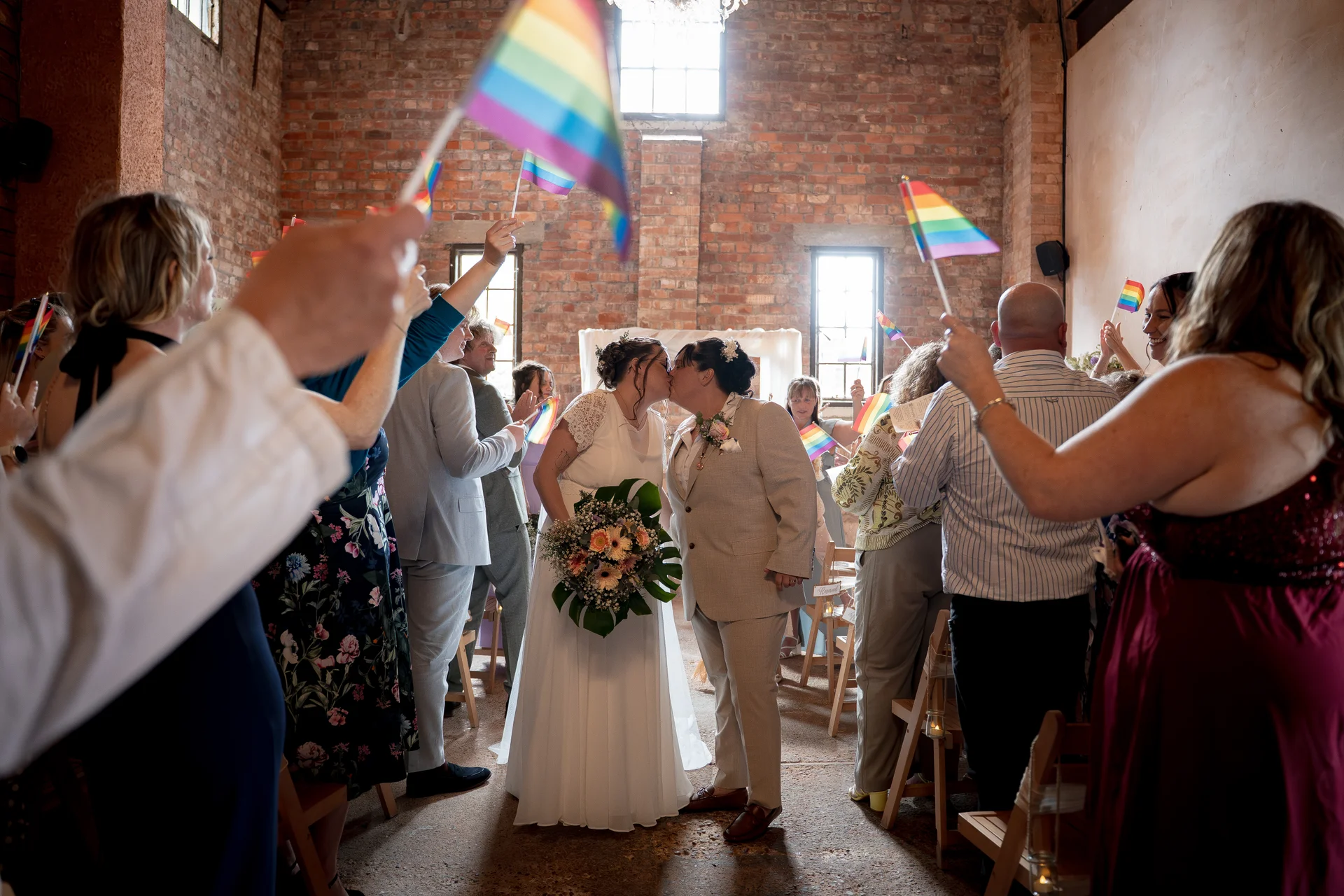A same-sex couple share a loving kiss as they walk down the aisle in a rustic brick barn, surrounded by cheering guests waving rainbow flags.