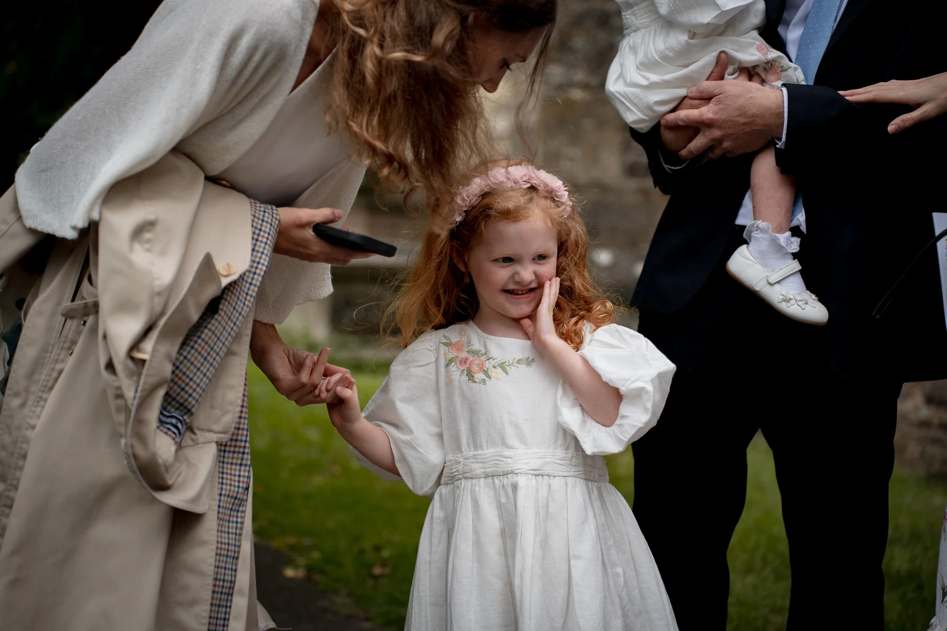 A young flower girl with red curly hair and a floral crown smiles shyly while holding hands with a guest outdoors during a wedding.