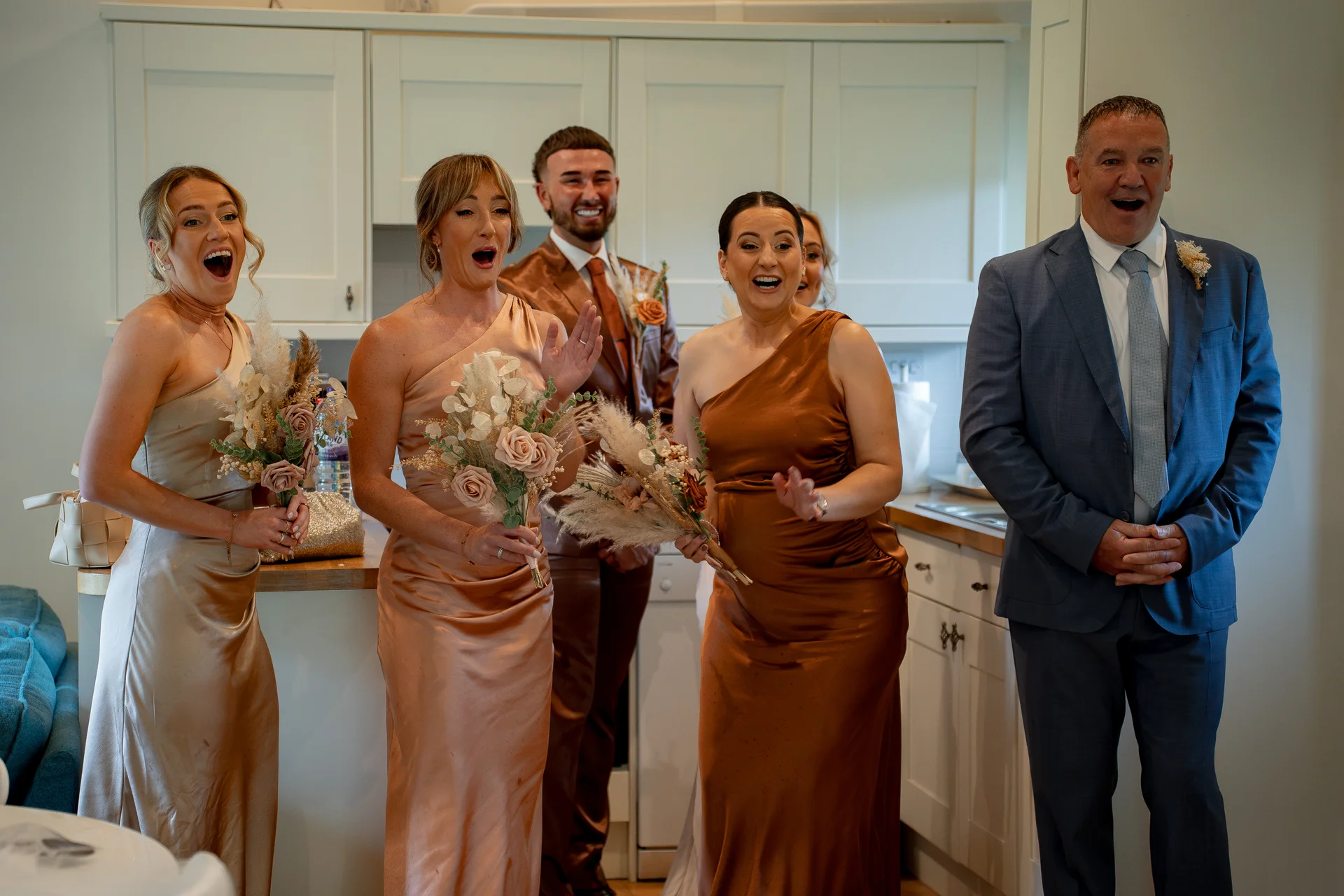 A group of delighted bridesmaids in satin dresses and a groomsman express joyful surprise in a bright kitchen interior.