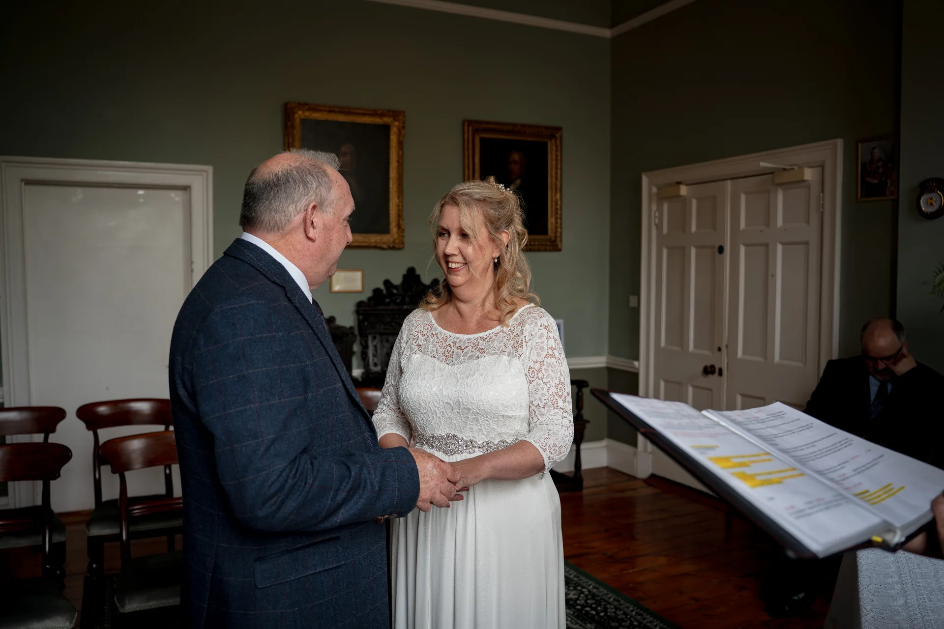 An older couple holds hands and smiles during an indoor civil ceremony inside a room with panelled green walls and framed portraits.