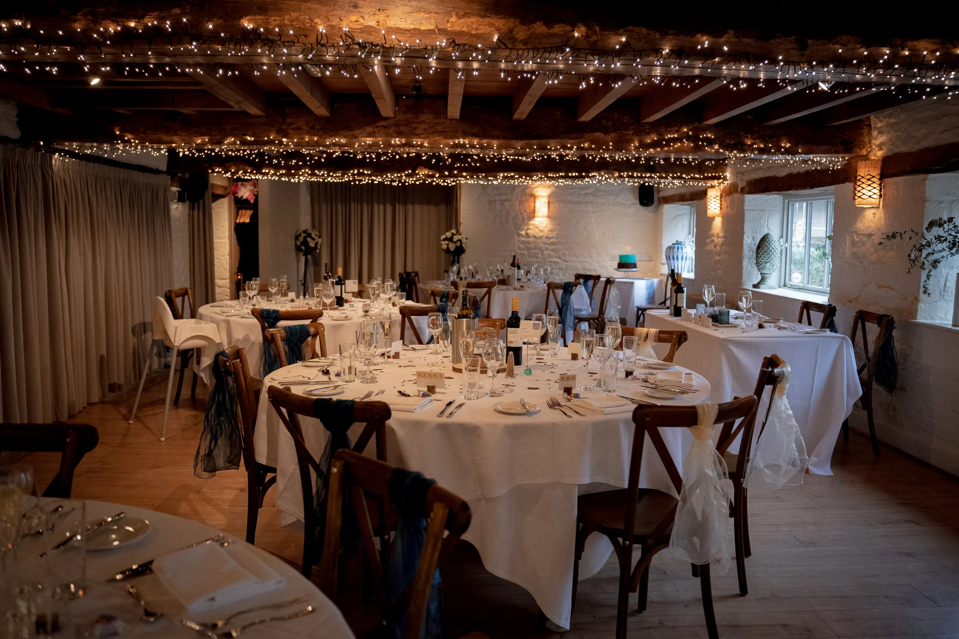 A rustic barn wedding reception room featuring round tables set with white linens, wooden chairs draped with sheer fabric, and an intricate canopy of fairy lights strung across exposed ceiling beams.