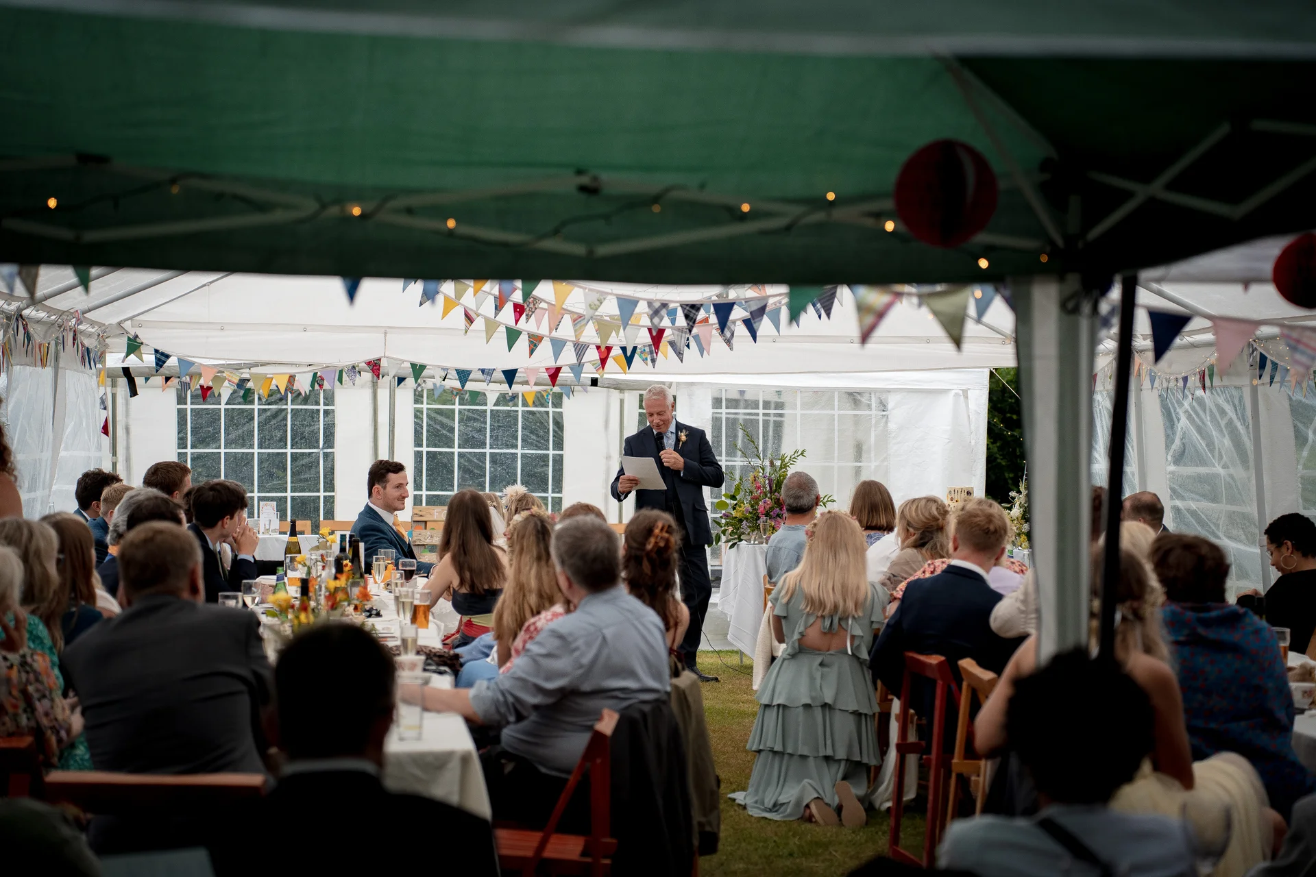 A guest delivers a speech during the wedding breakfast inside a brightly lit marquee adorned with colourful bunting.