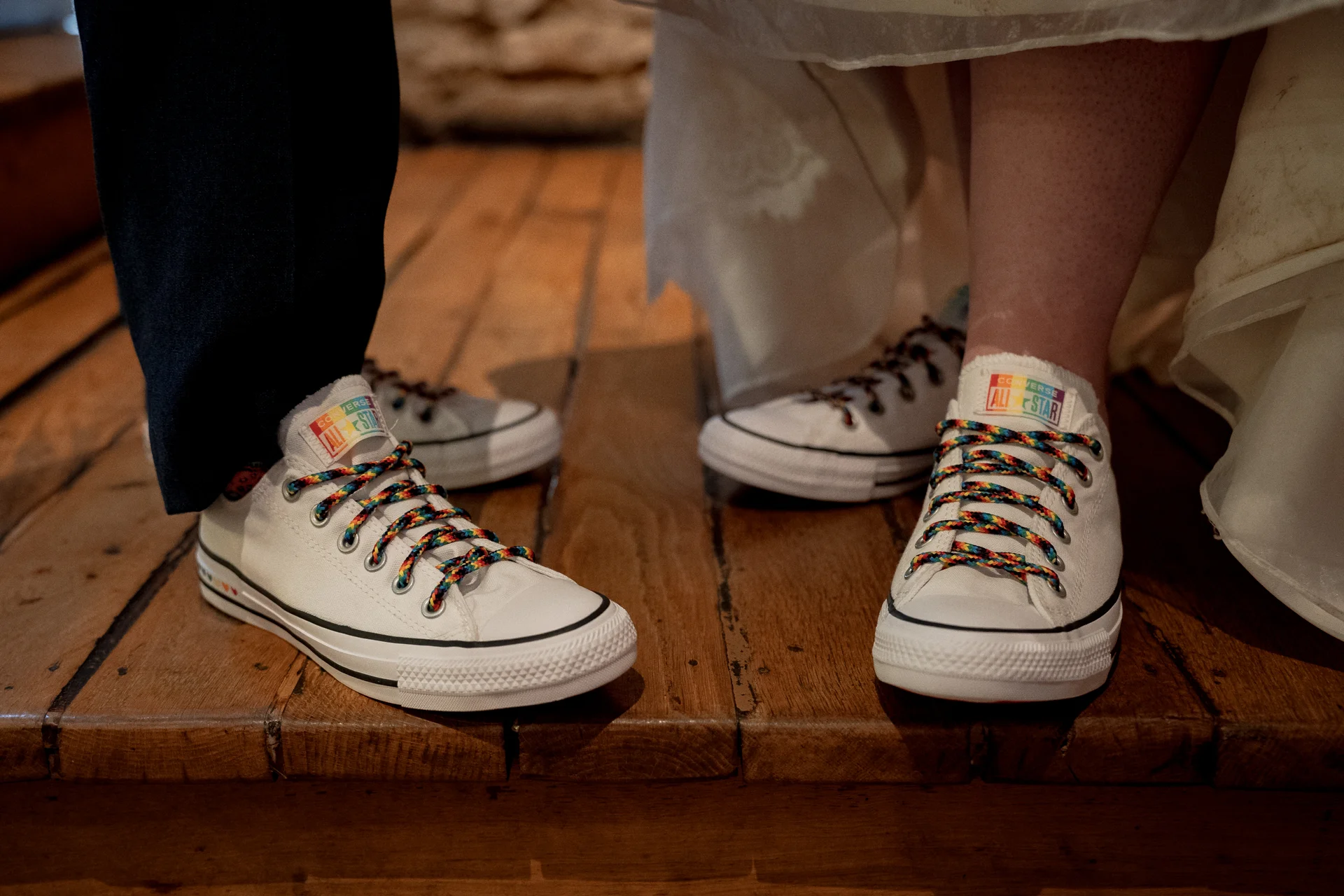 Close-up of a newlywed couple wearing matching white Converse trainers with colourful rainbow laces while standing on a rustic wooden floor.