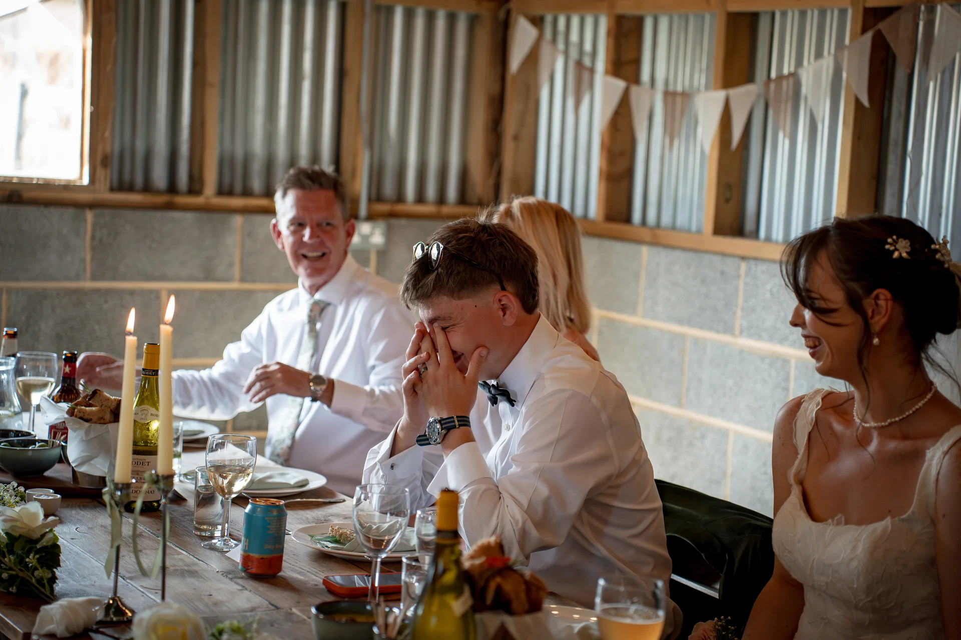 A groom covers his face with laughter during wedding speeches at a long banquet table inside a rustic barn venue decorated with bunting and candles.