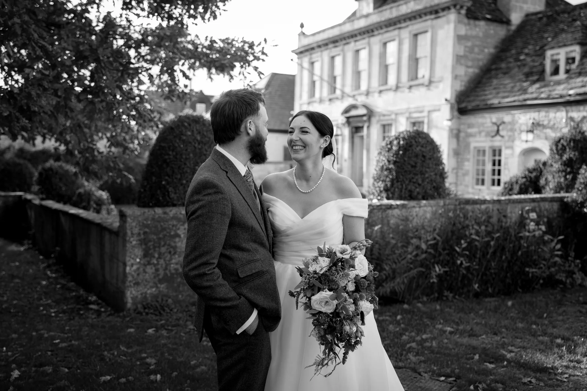 A newlywed couple share a joyful glance in the manicured gardens of a classic country house, with the bride holding a cascading bouquet.