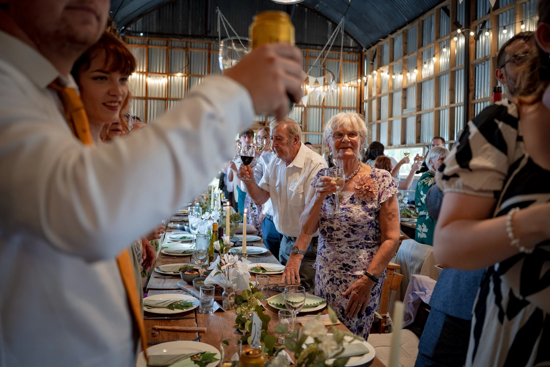 Wedding guests raise their glasses for a toast during a lively wedding breakfast inside a rustic barn with exposed rafters and string lights.
