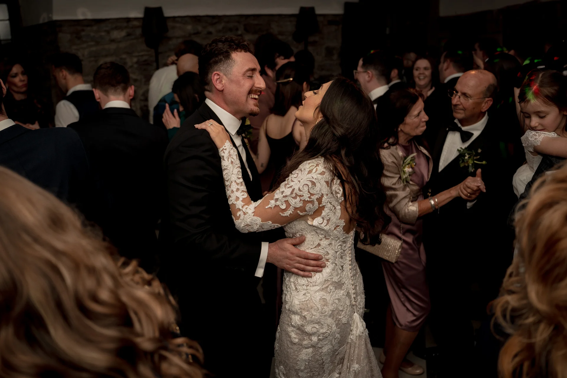 A newlywed couple shares a joyful first dance surrounded by family and guests on a crowded evening reception dance floor.