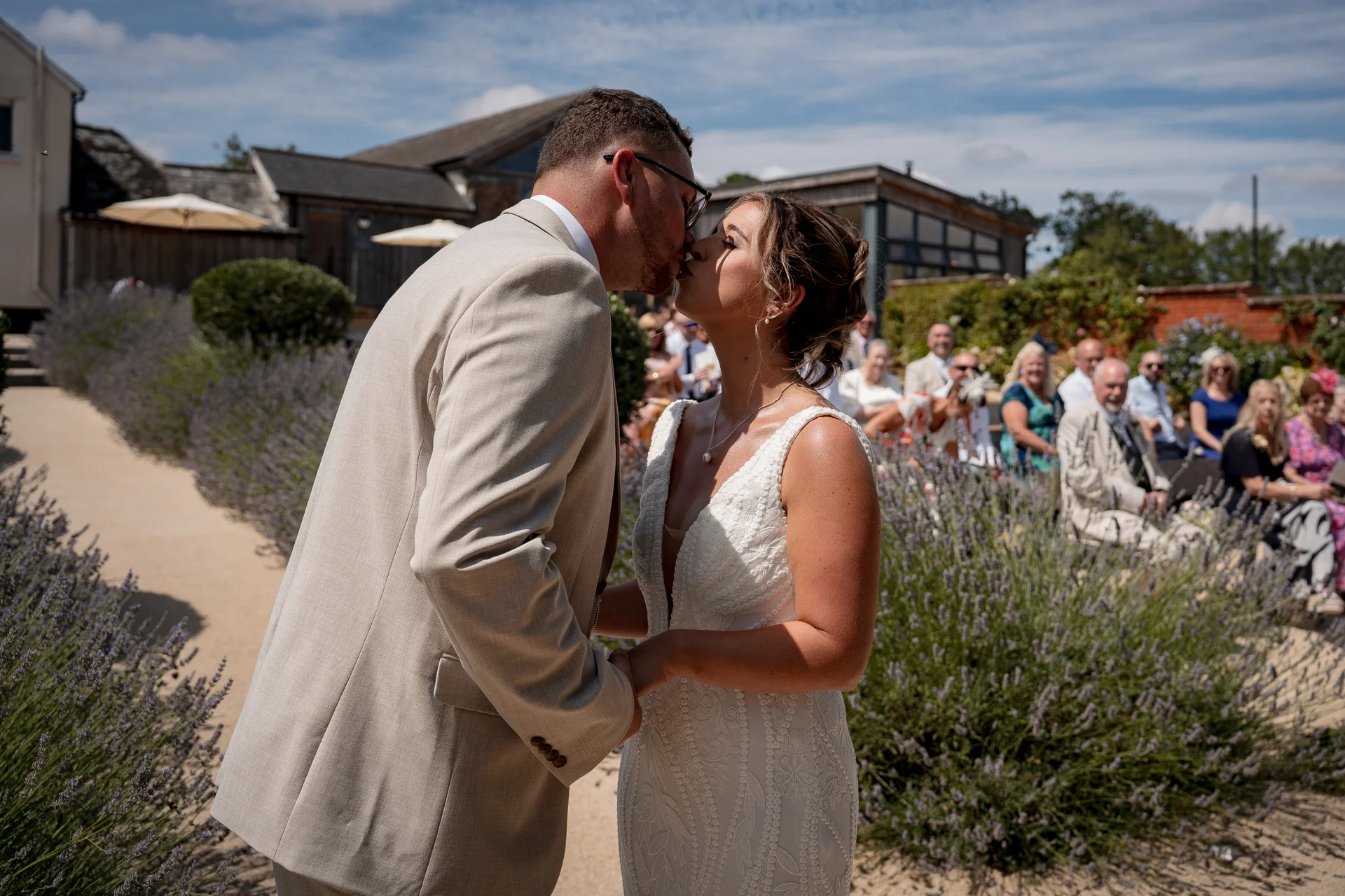 Newlywed bride and groom share their first kiss during an outdoor summer ceremony surrounded by lavender beds and seated guests.
