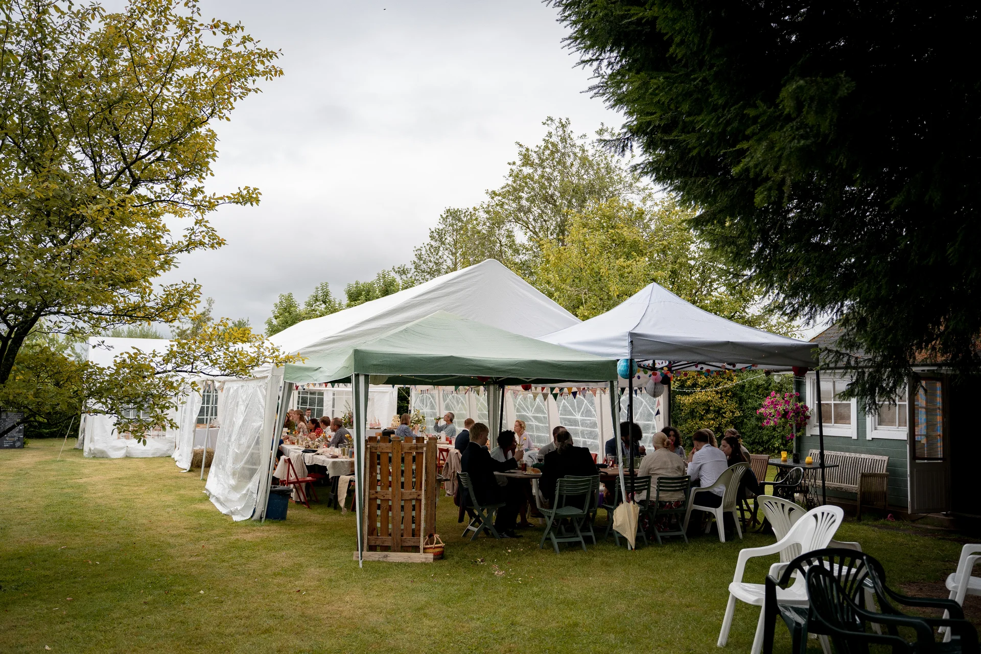 Guests enjoy a wedding breakfast seated beneath connected white and green marquees on an open lawn surrounded by mature trees.
