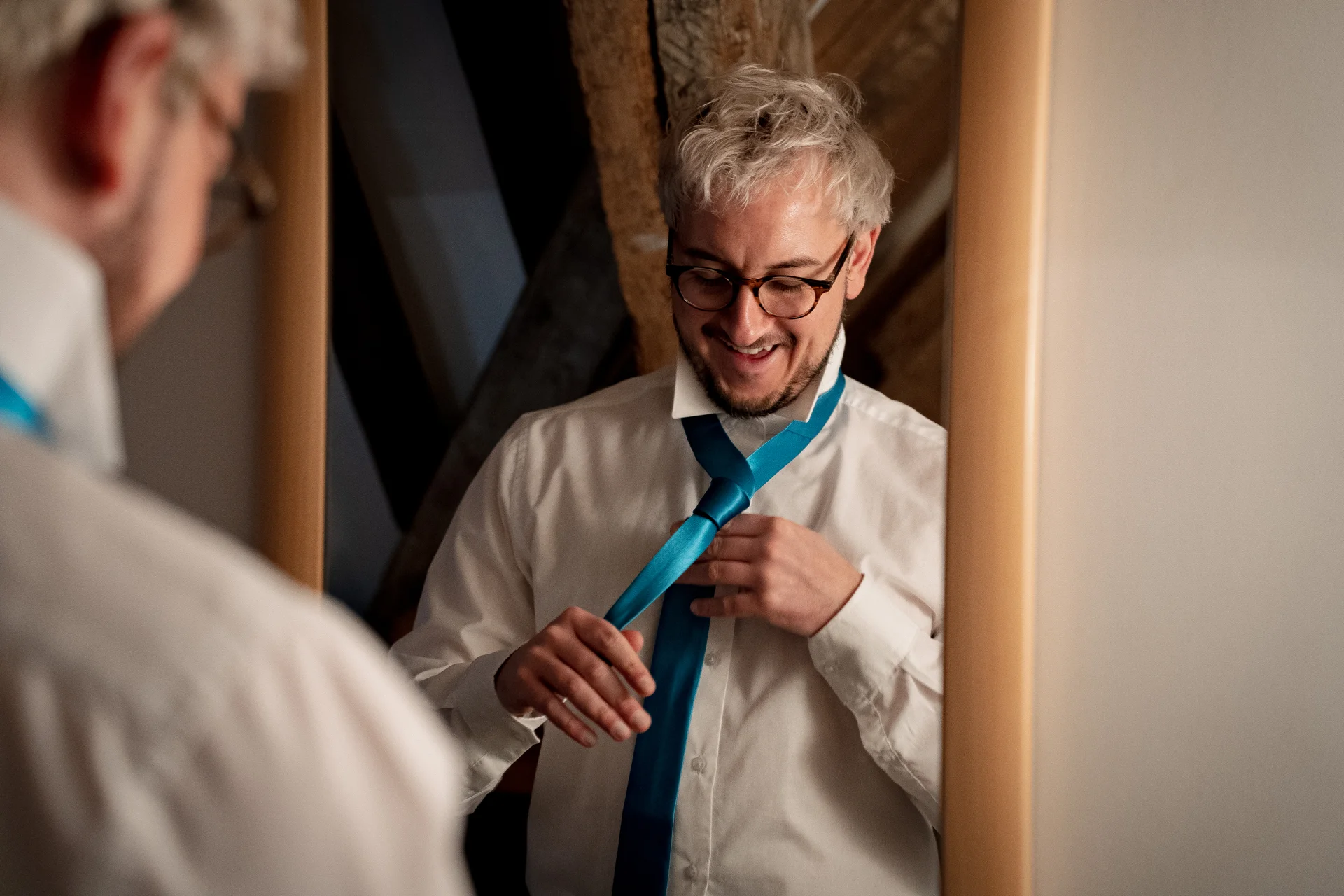 A smiling groom looks into a mirror while tying his vibrant blue tie during morning wedding preparations.