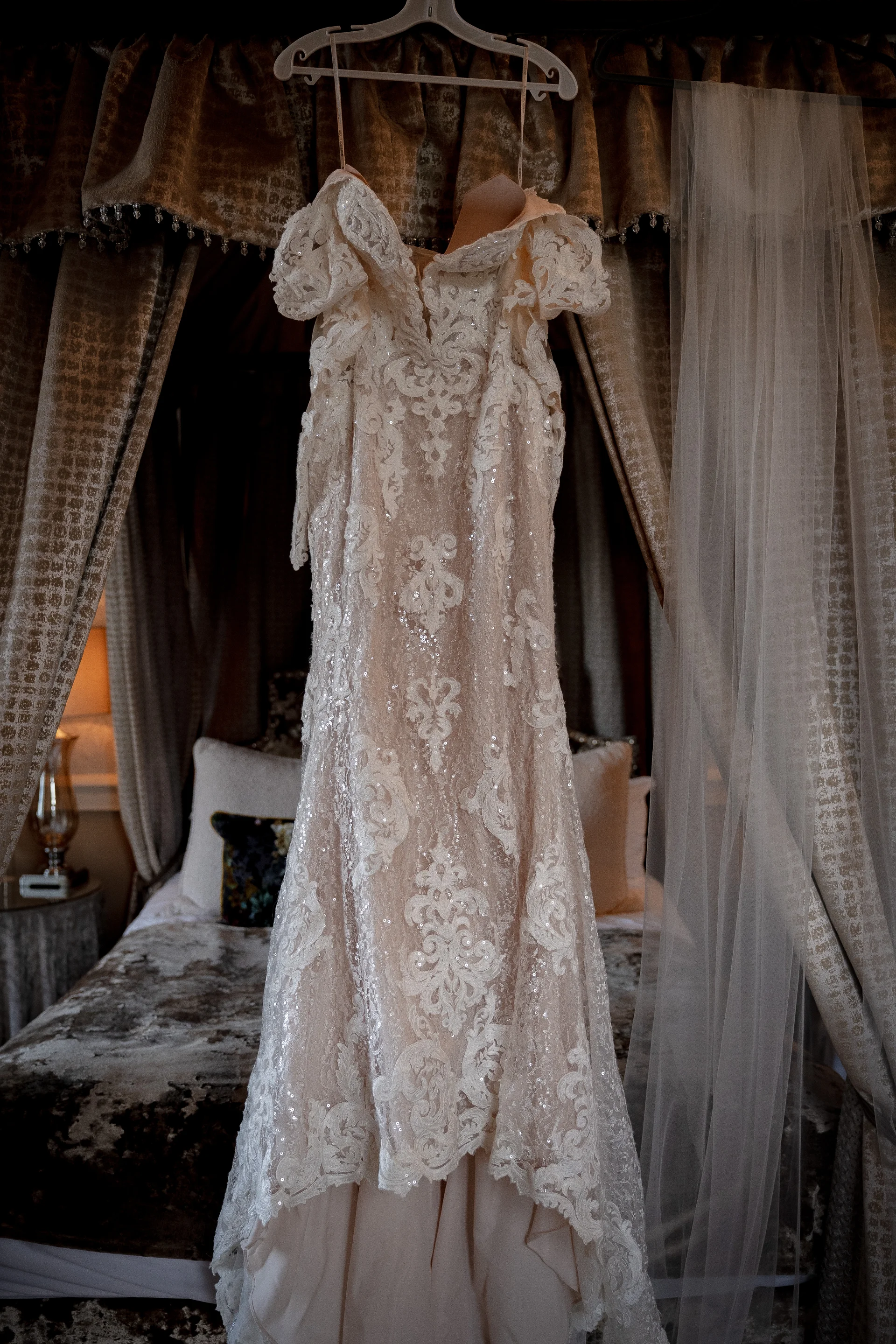 An intricately beaded lace wedding dress with off-the-shoulder straps hangs against a draped canopy bed in a moody bedroom setting.