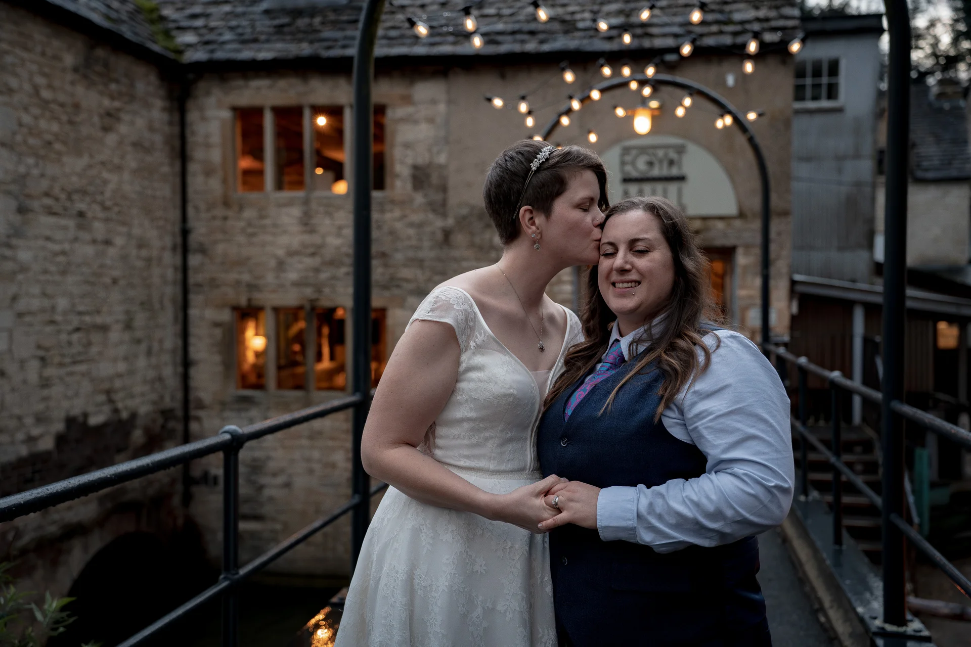 A newlywed couple on a stone bridge at twilight, with one bride giving the other a tender forehead kiss beneath a glowing arch of festoon lights.