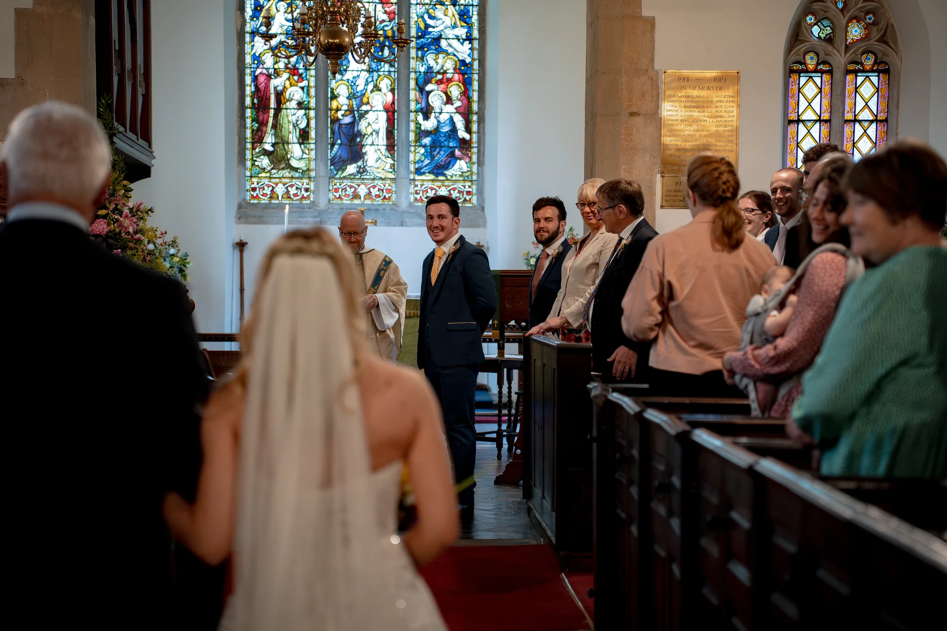 The bride walks down the aisle in a traditional church while the smiling groom watches from the altar.