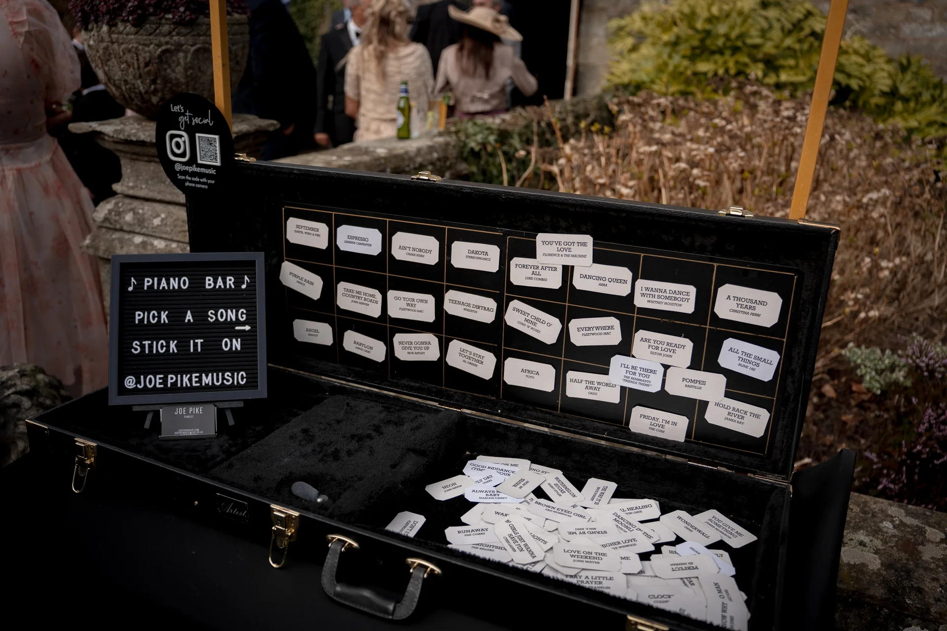 An open black instrument case filled with song title tags and a felt letter board invites wedding guests to pick and stick their favourite requests for the piano bar.