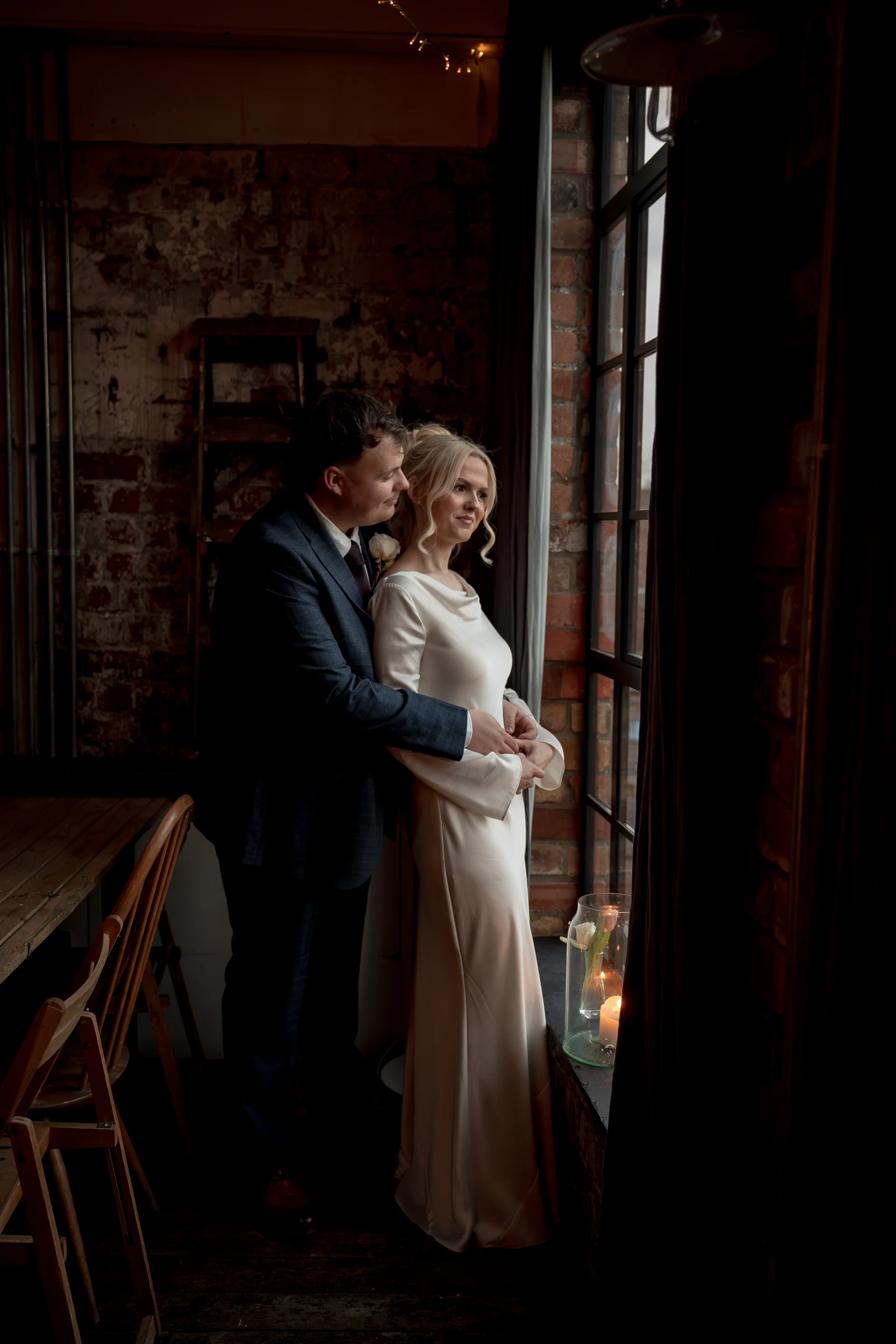 A newlywed couple embraces tenderly by a large industrial window inside a rustic brick venue, illuminated by soft natural light and glowing candles.