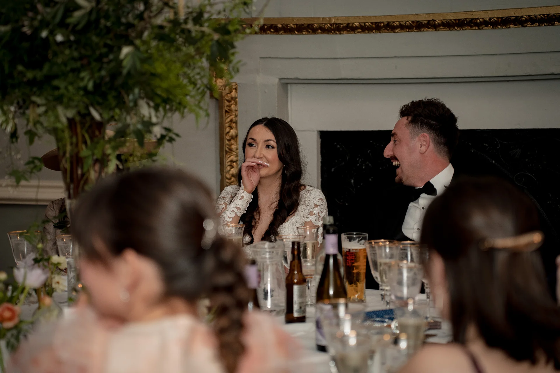 A newlywed couple sits at the top table during wedding speeches, laughing together in a candlelit room framed by lush floral arrangements.