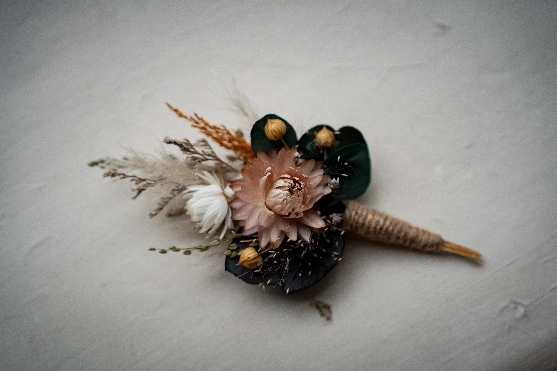 A detailed flat lay shot of a rustic dried flower buttonhole featuring neutral blooms, seed pods, and twine wrapping on a pale surface.