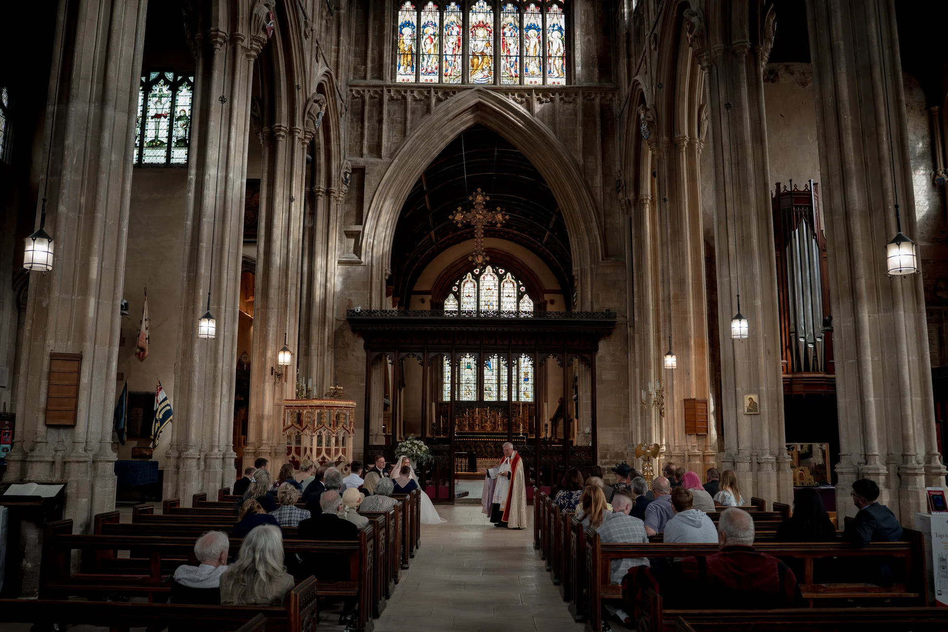 A wide-angle interior view of a grand stone cathedral during a wedding ceremony, showing seated guests in wooden pews facing the altar under soaring stained glass windows and vaulted ceilings.