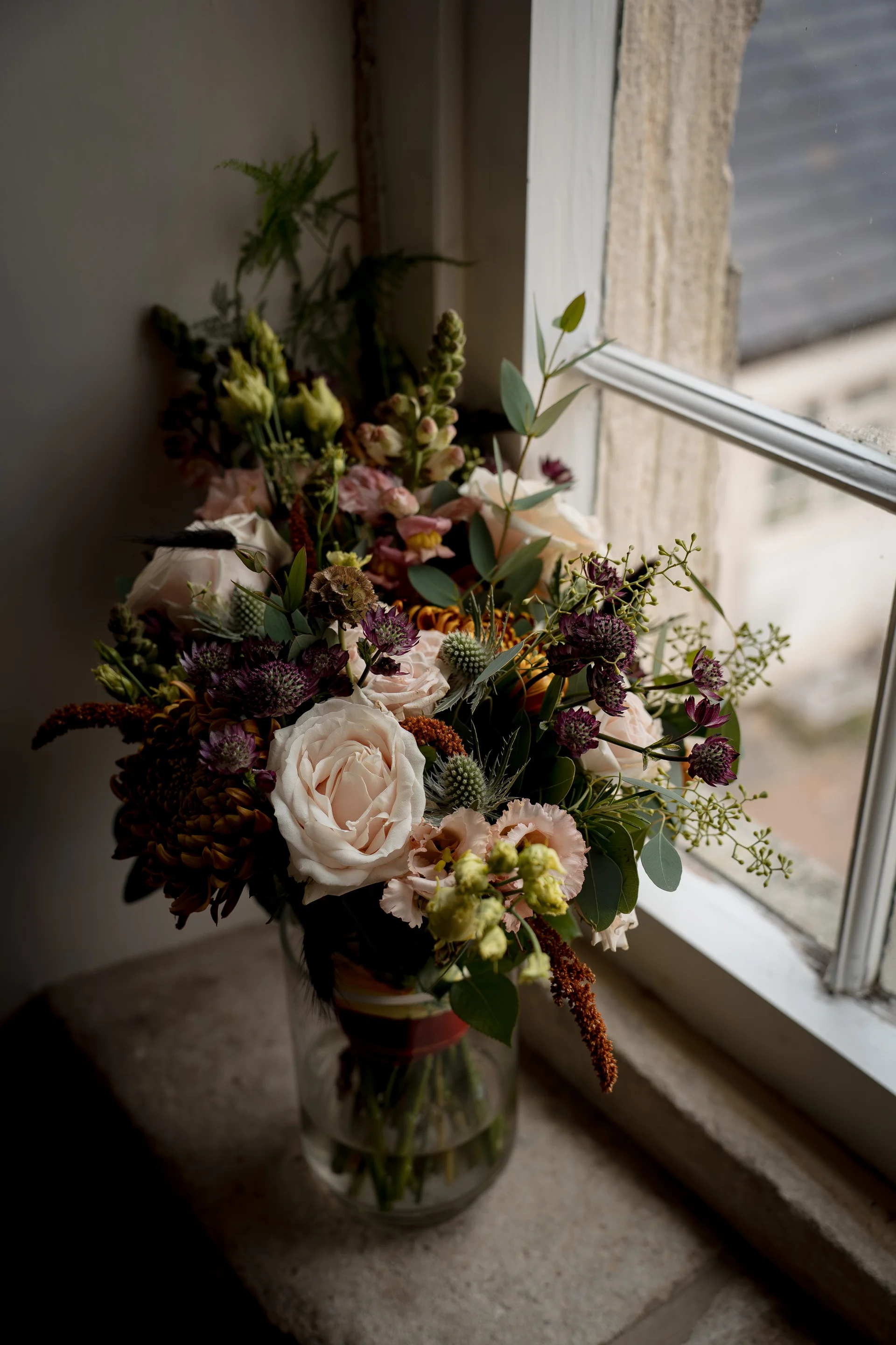 A lush, hand-tied autumn wedding bouquet rests in a clear glass vase on a stone windowsill, illuminated by soft natural window light.