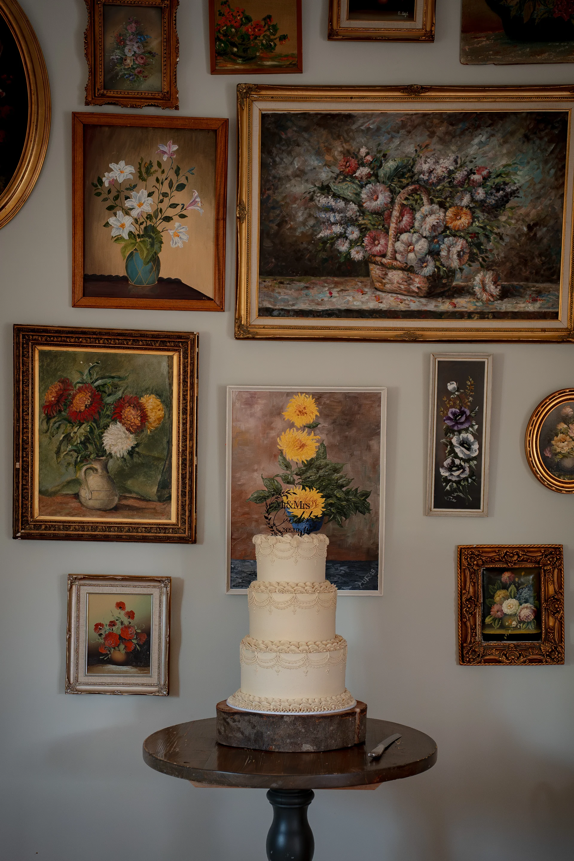 A three-tier white buttercream wedding cake with intricate piping sits on a wooden slice table against a gallery wall filled with vintage floral paintings.