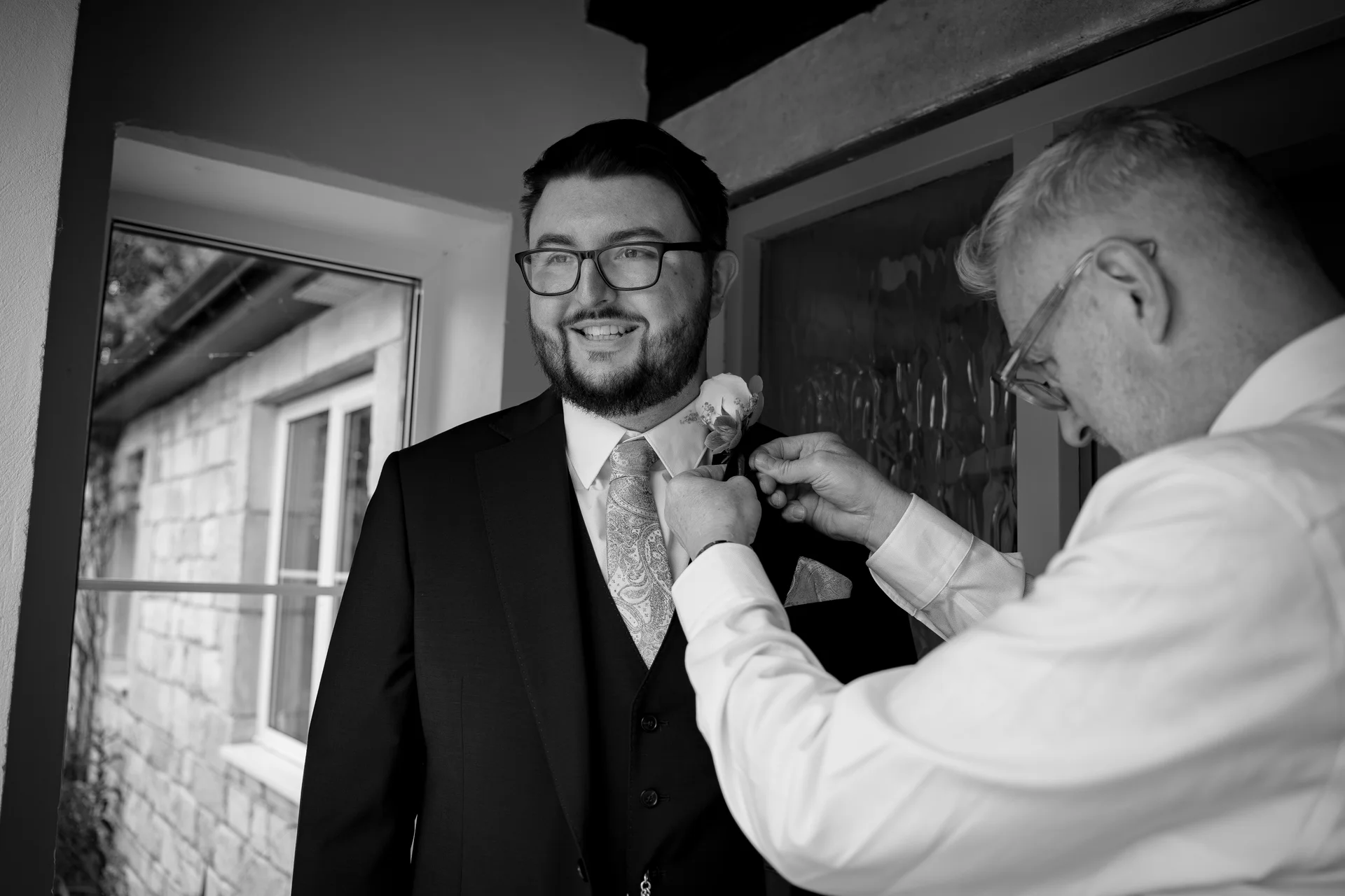 A smiling groom wearing a three-piece suit has his boutonniere pinned to his lapel by another man during morning preparations.