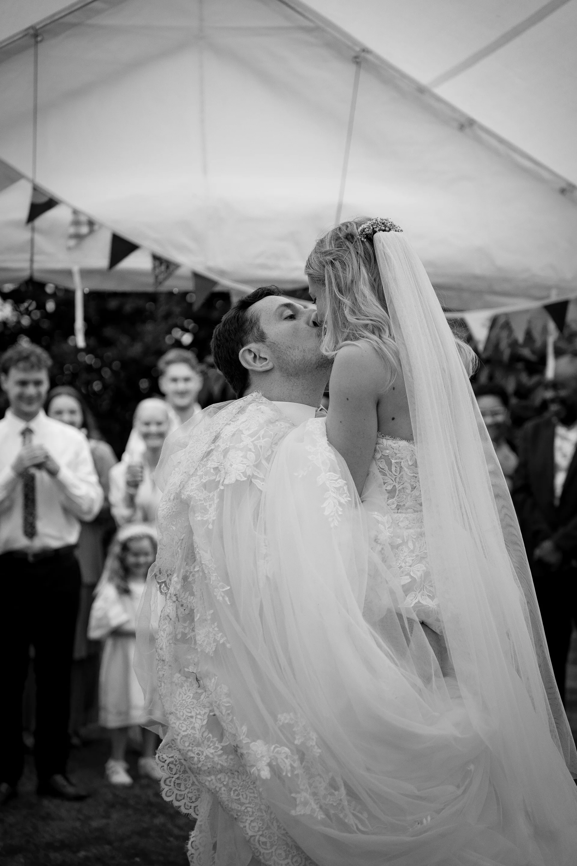 A groom joyfully lifts and kisses his bride under a white marquee, surrounded by cheering wedding guests and classic bunting.