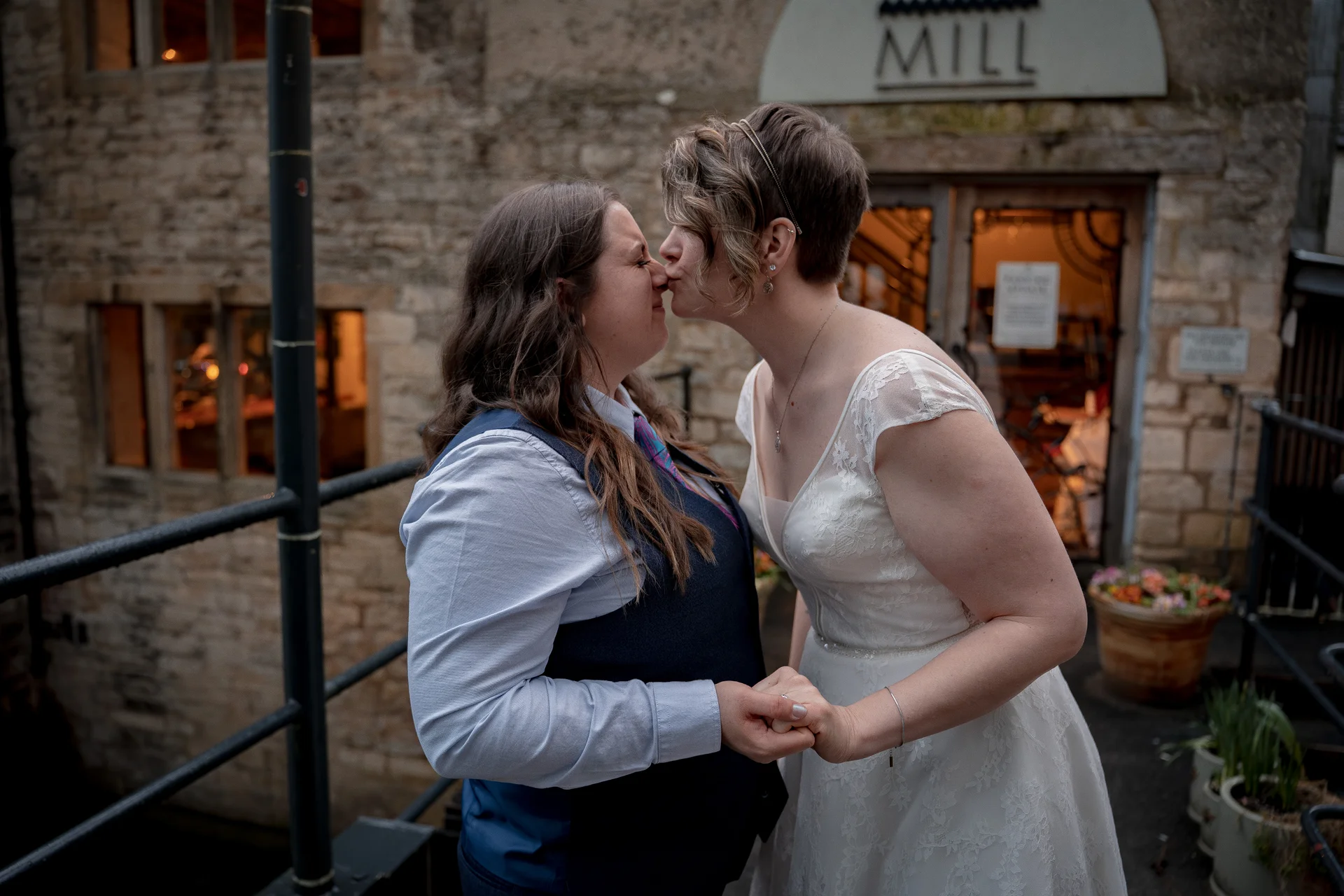 Two brides share a tender nose-to-nose kiss outside a rustic stone building, holding hands while dressed in a lace wedding dress and a smart waistcoat.