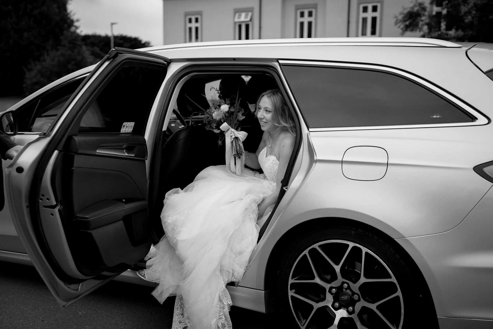 A smiling bride wearing a strapless lace gown sits inside the open door of a wedding car while holding a bouquet.