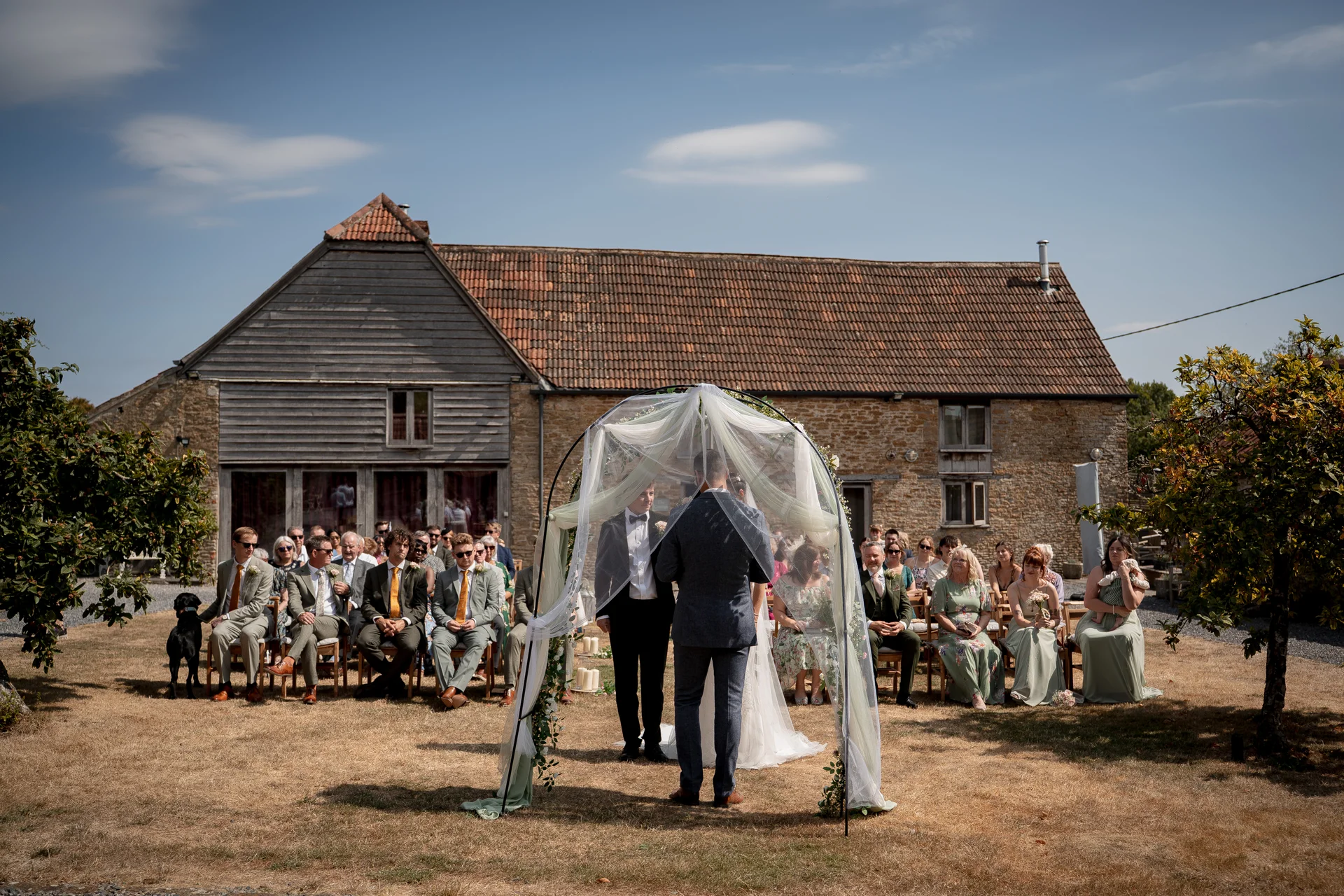 An outdoor wedding ceremony takes place in front of a rustic stone barn, with the couple standing beneath a fabric-draped arch before seated guests under a bright blue summer sky.