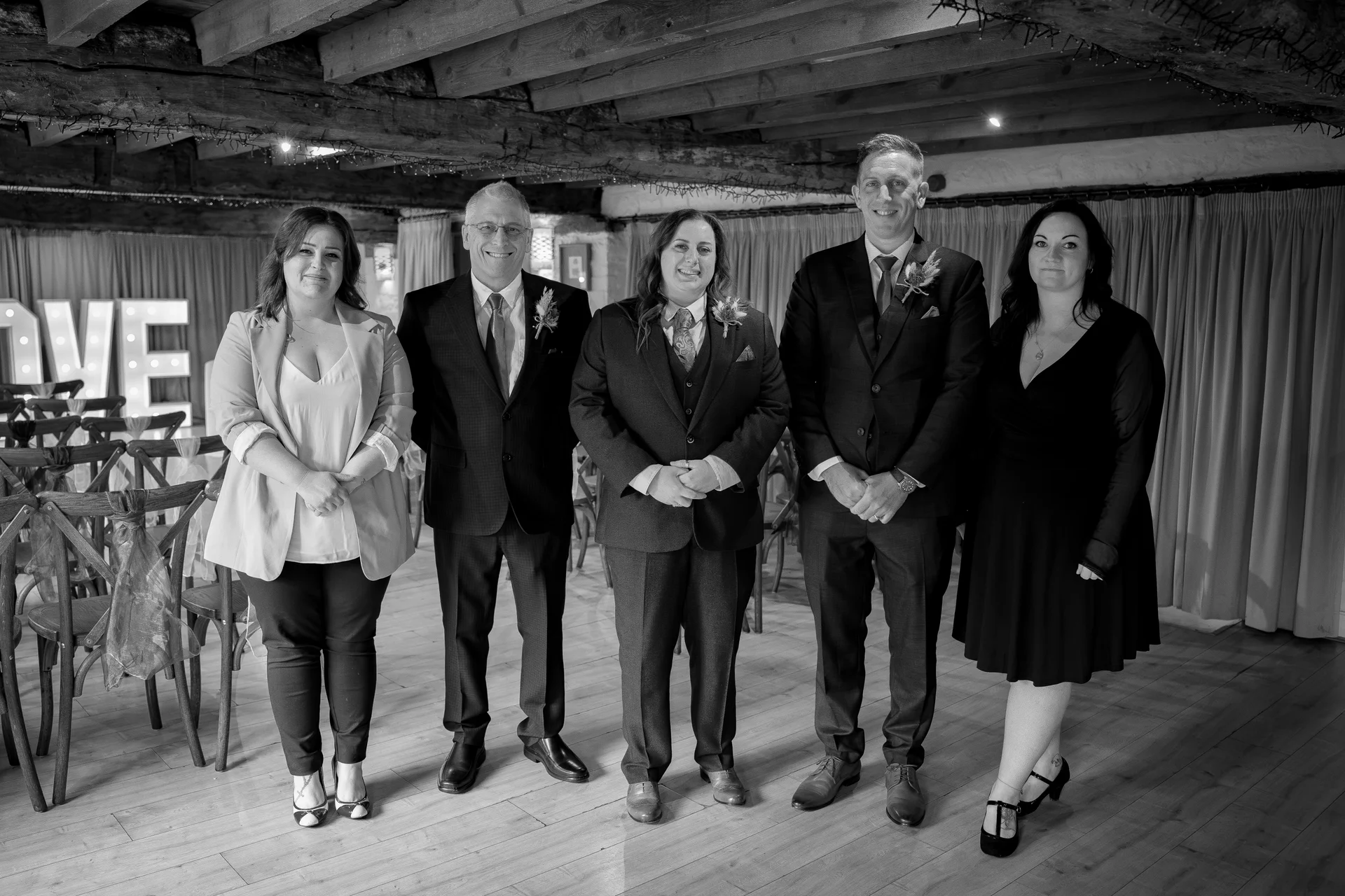 A group of five wedding guests posing together inside a rustic barn with exposed wooden rafters and wooden flooring.