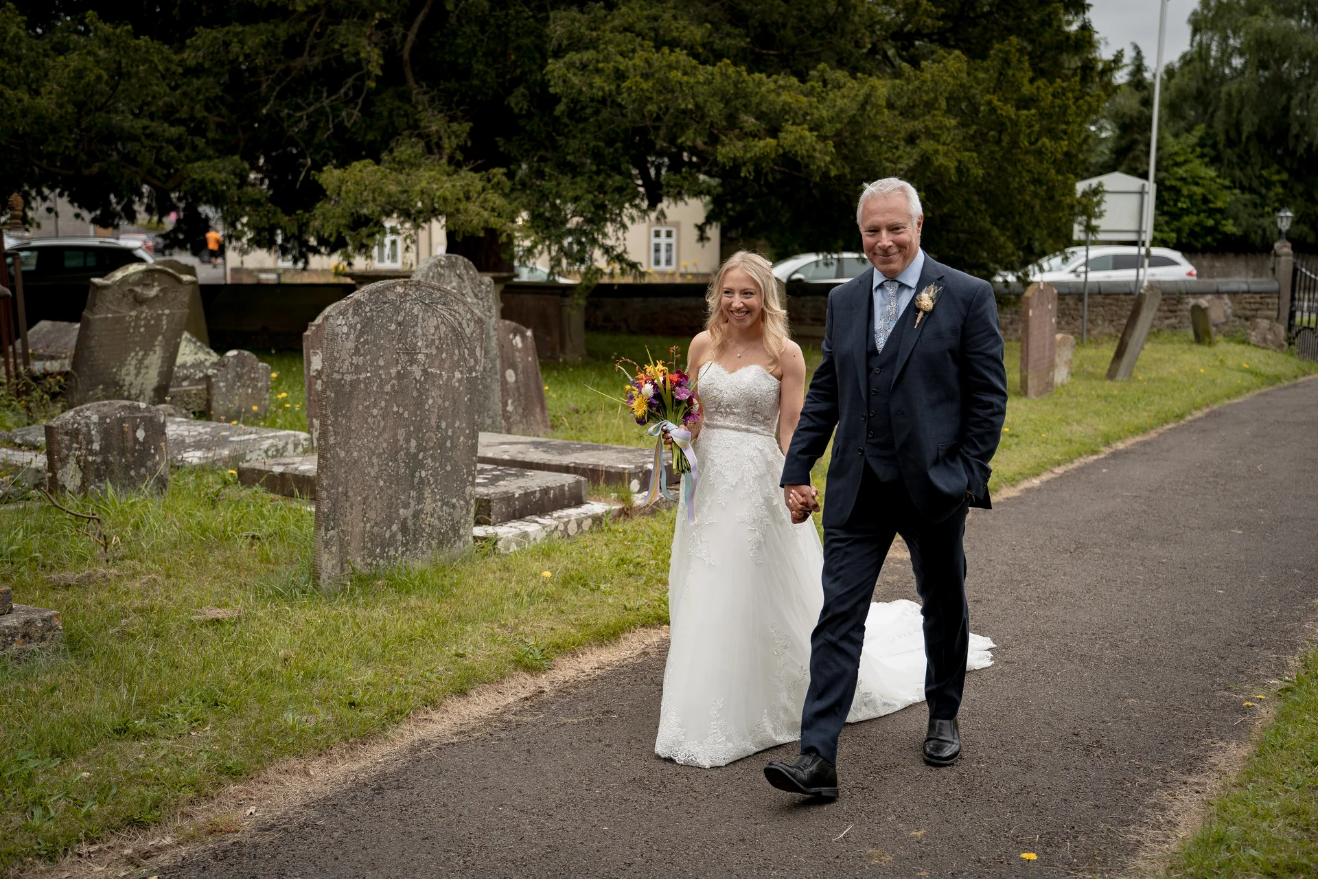 A smiling bride holding a colourful wildflower bouquet walks hand in hand with her father down a path beside a churchyard.