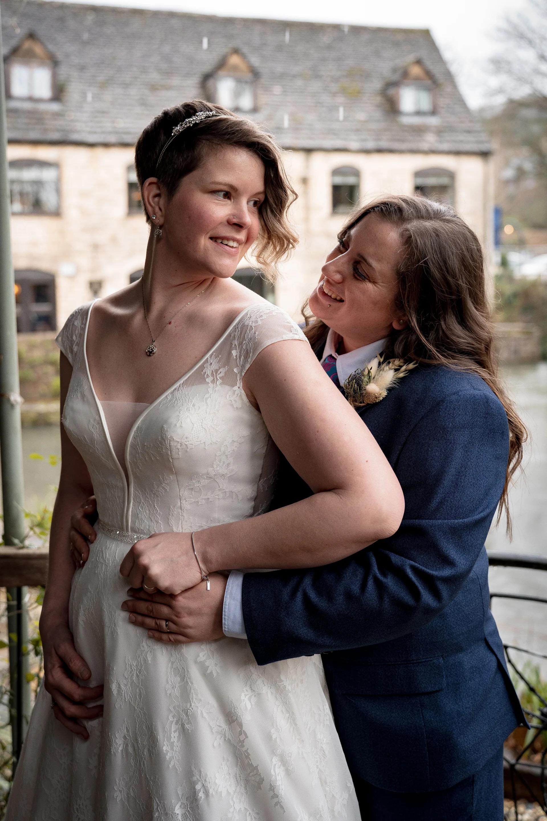 A happy same-sex couple embracing outdoors, with one bride wearing a lace wedding dress and the other wearing a smart navy suit.