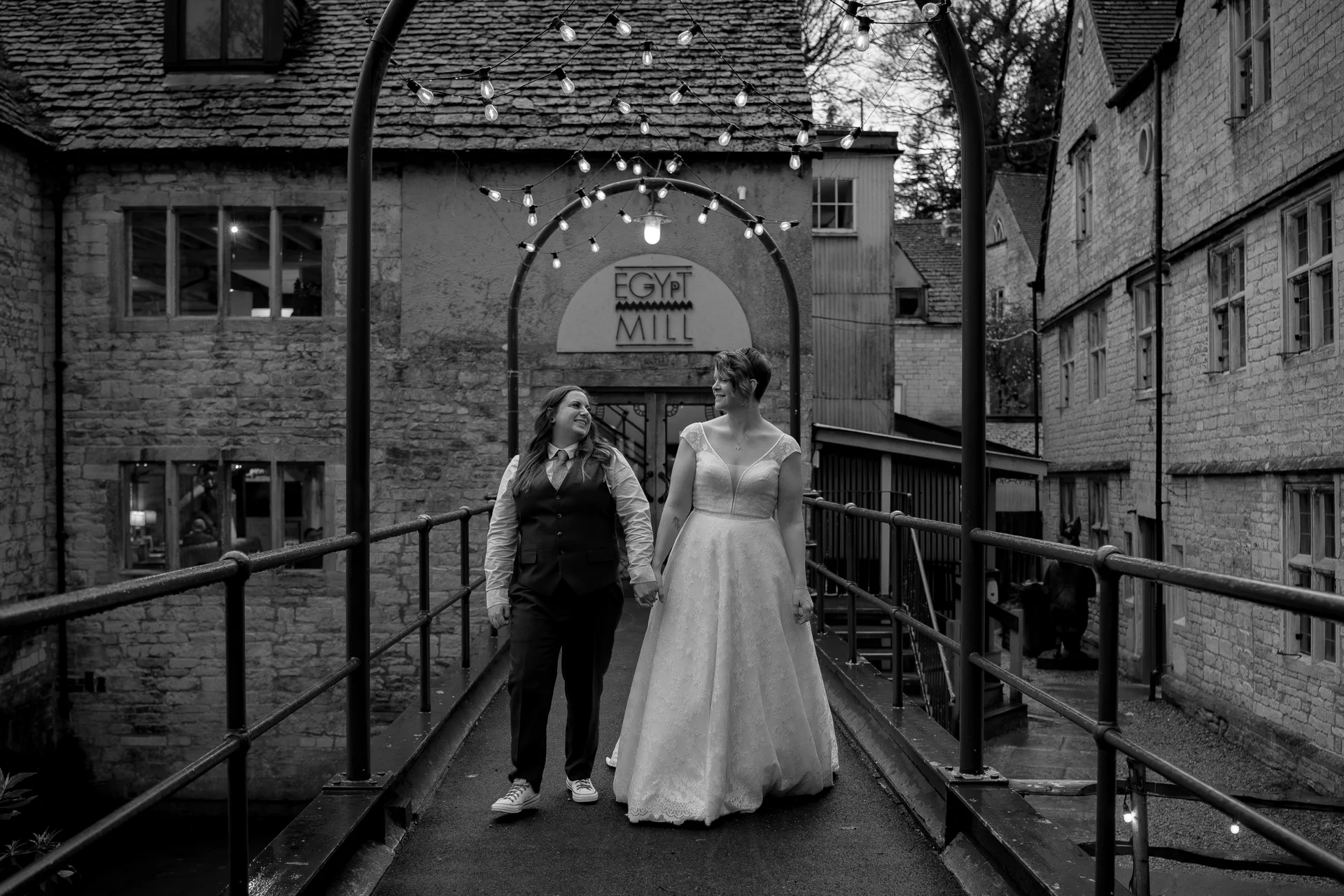 A newlywed same-sex couple lovingly gaze at each other while walking hand-in-hand across a stone bridge framed by rustic buildings and sparkling festoon lights.