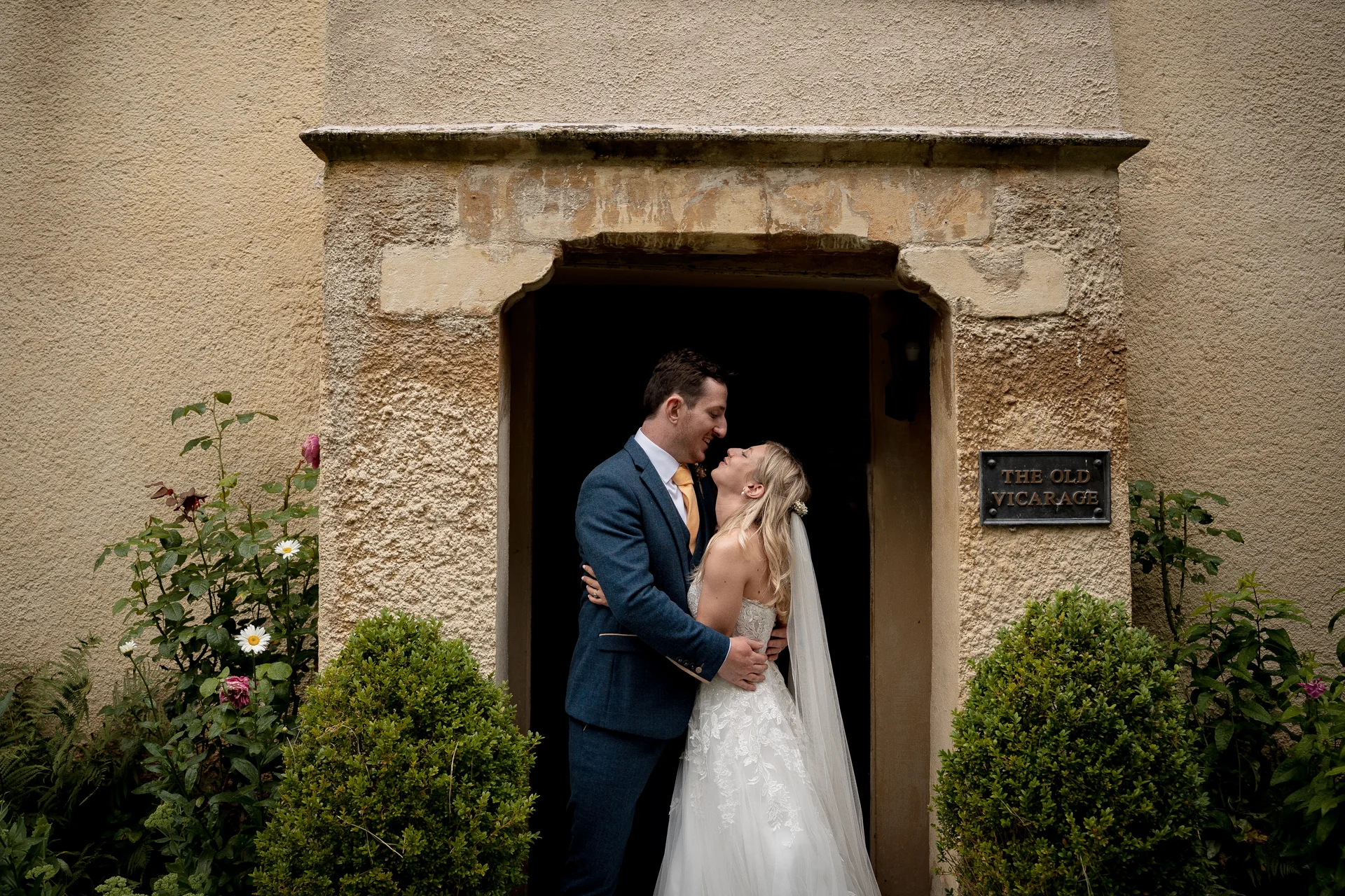 A newlywed couple embraces lovingly within the rustic stone entrance doorway of a historic country venue, framed by lush green foliage and neatly trimmed boxwood shrubs.