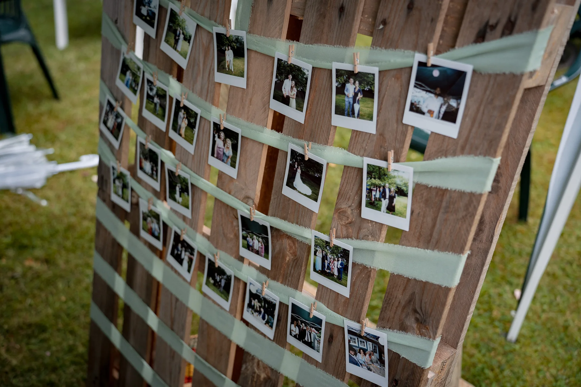 A rustic wooden pallet holding numerous polaroid style photographs pinned with small pegs and sage green ribbons on a lawn.
