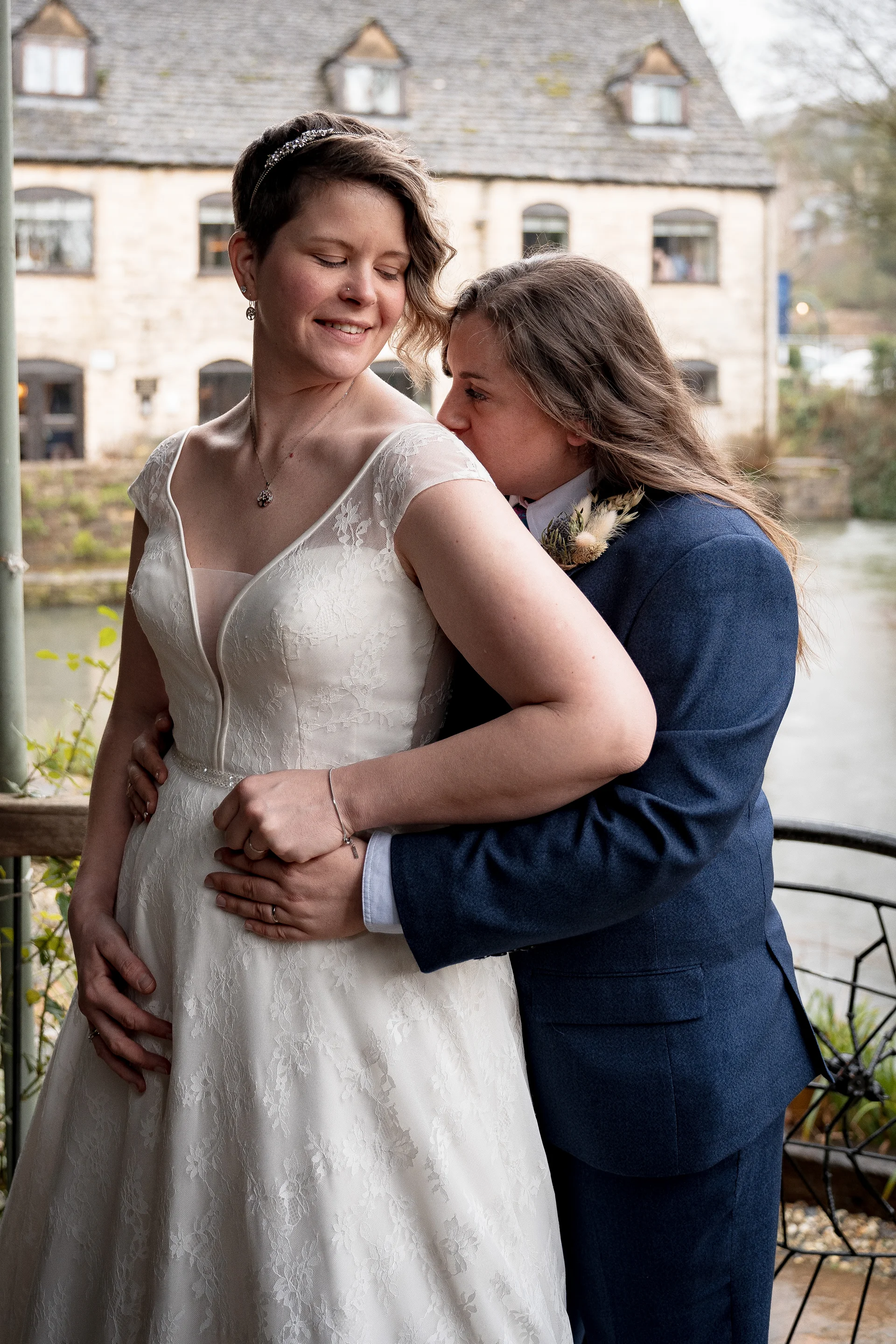 Two brides embrace lovingly outdoors with a historic stone building in the background, one wearing a lace wedding dress and the other in a navy suit.
