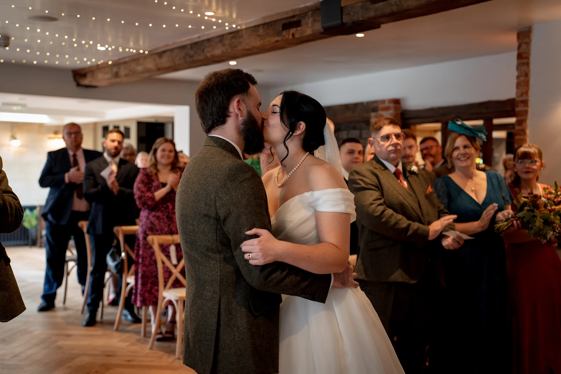 Newlywed bride and groom share their first kiss at the altar inside a rustic barn venue decorated with exposed wooden beams and fairy lights, as family and friends applaud in the background.