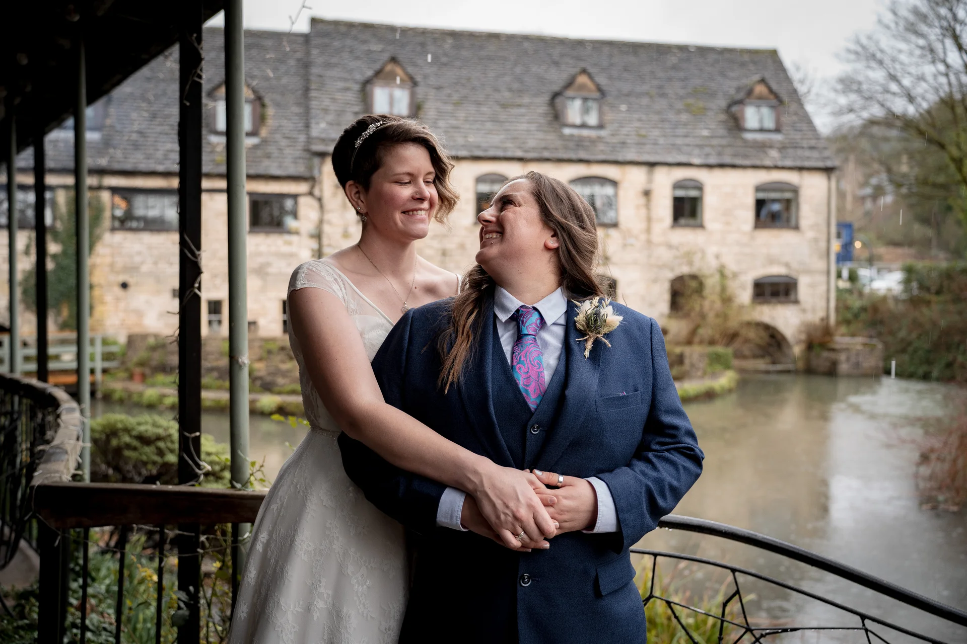 A same sex couple shares a tender embrace outside a rustic stone building by the water on their wedding day.
