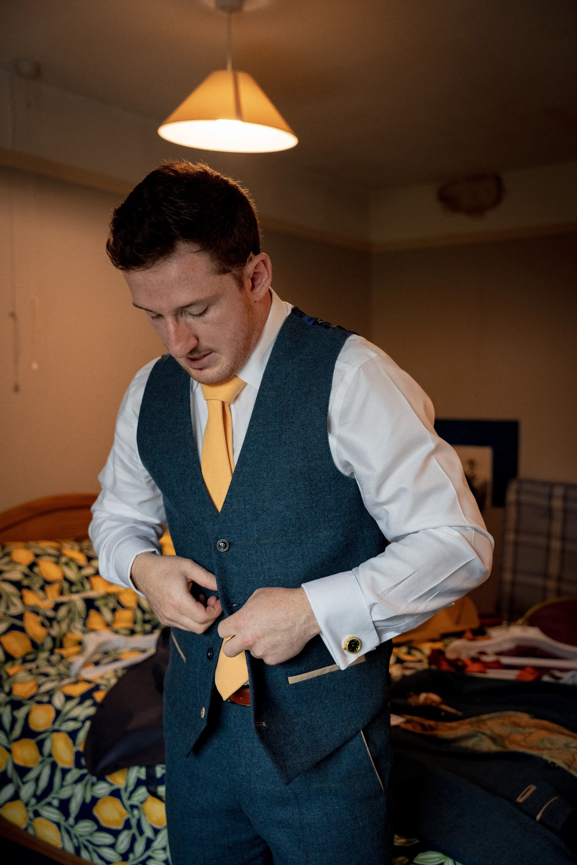 A groom in a blue tweed waistcoat and yellow tie carefully fastens his suit during morning preparations.