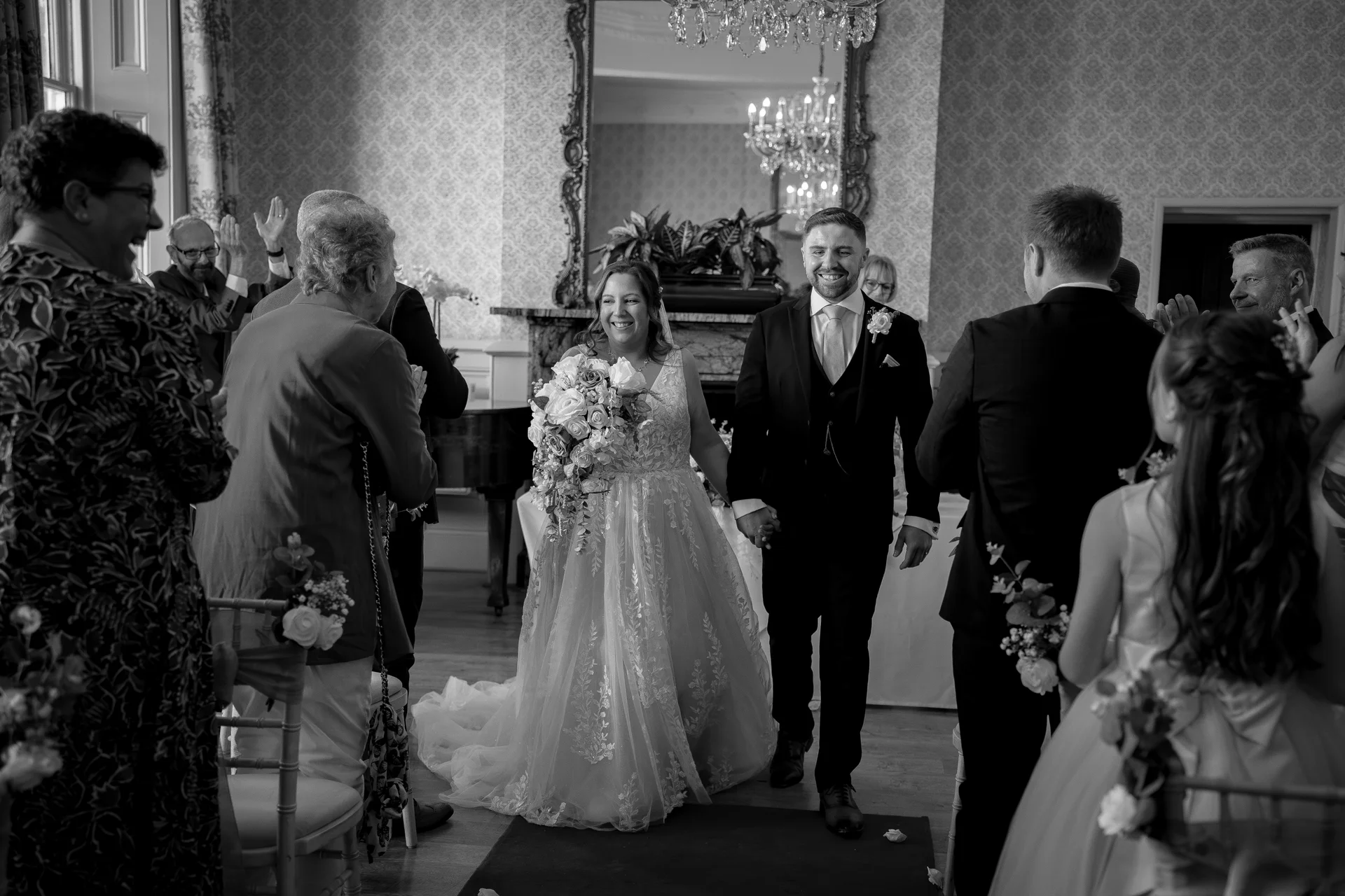 Newlyweds walk hand in hand down the aisle smiling joyfully as guests clap and cheer in a bright room.