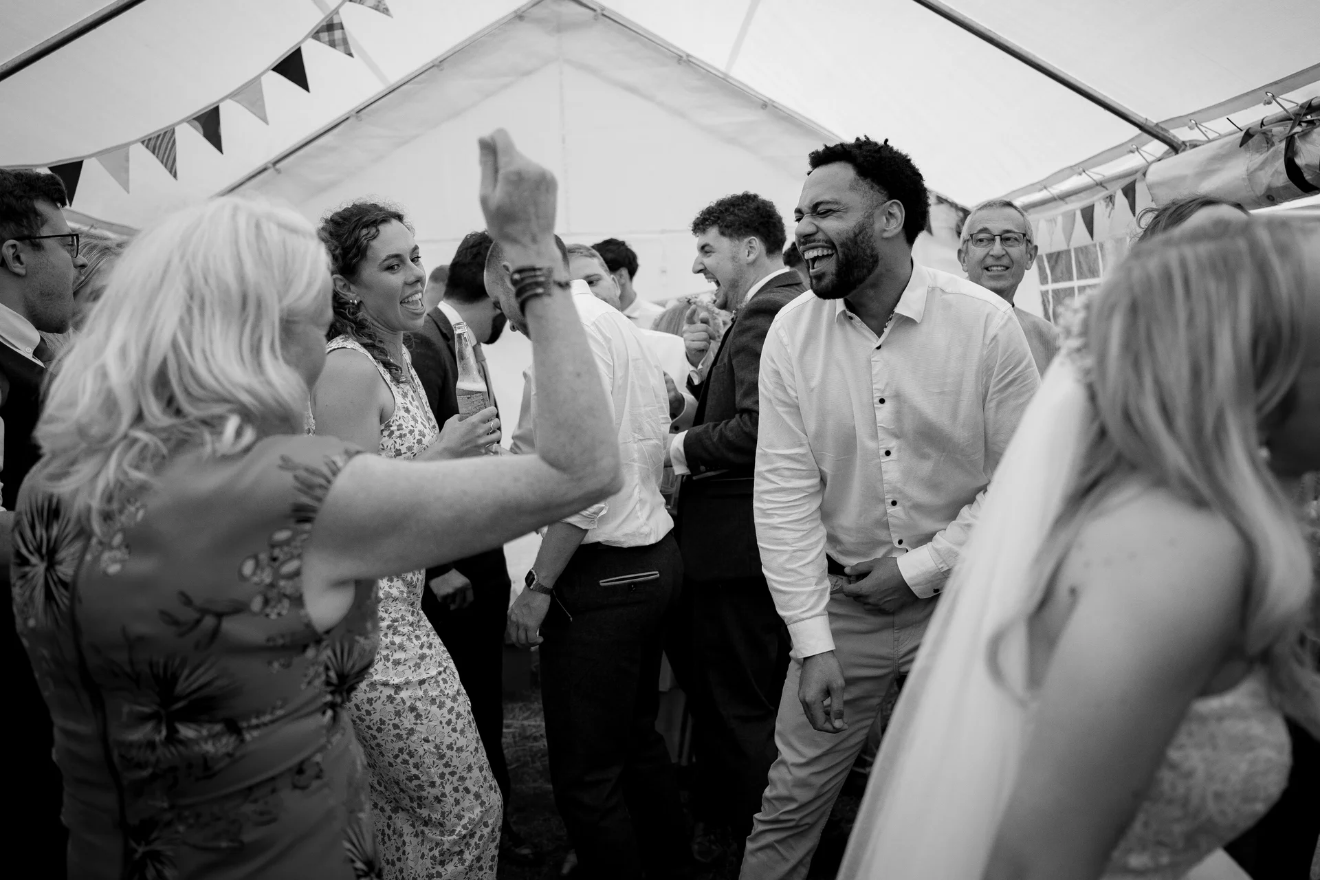 A candid black and white photograph captures wedding guests laughing and dancing together inside a marquee decorated with hanging bunting.