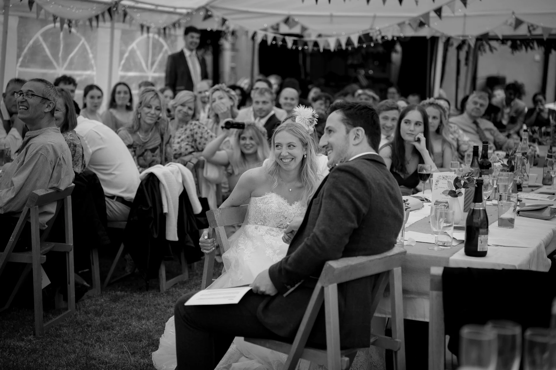 A bride and groom share a joyful laugh together seated at the top table during wedding speeches inside a marquee adorned with bunting.
