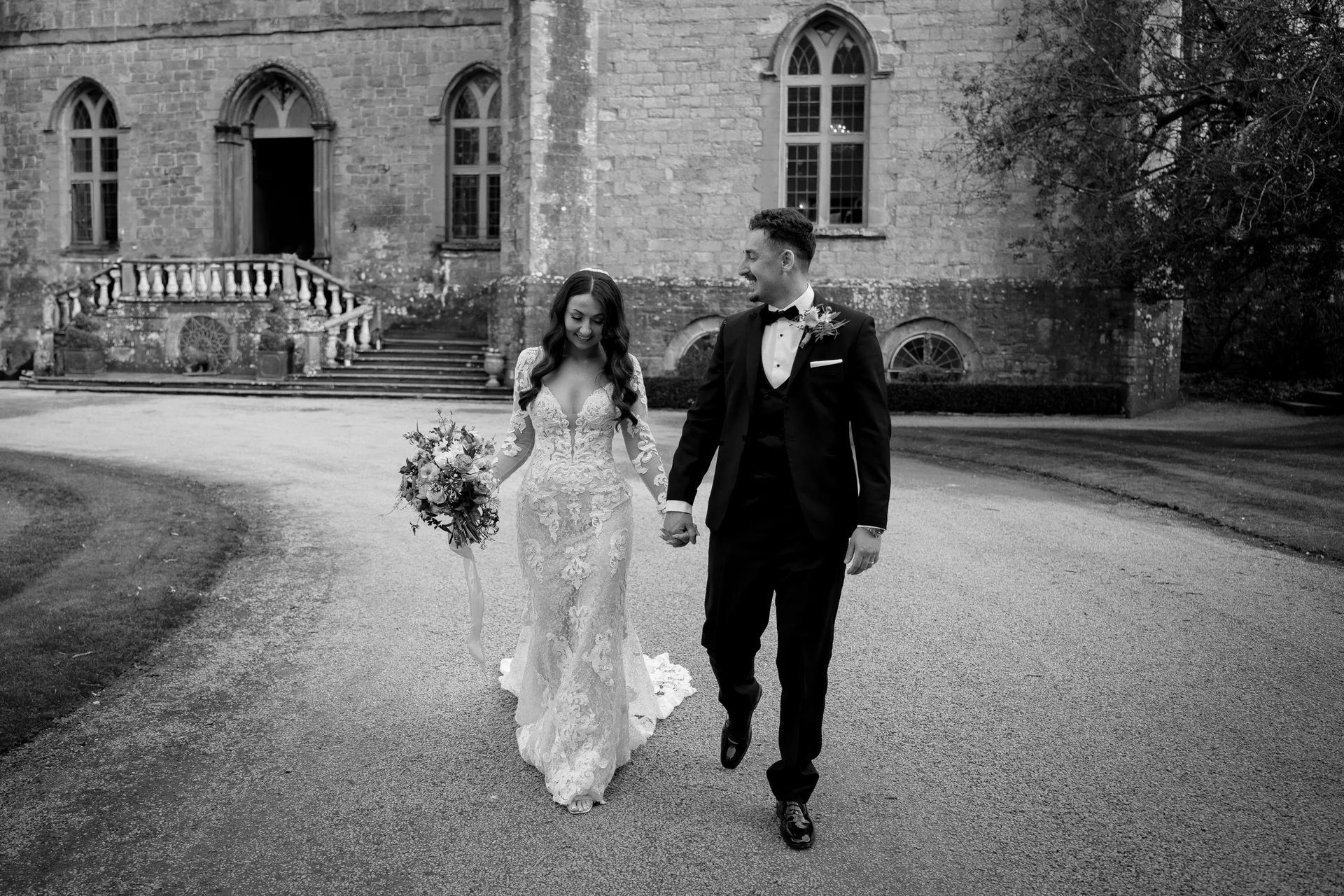 A newlywed couple holds hands and smiles at each other while walking outside a historic stone building on their wedding day.