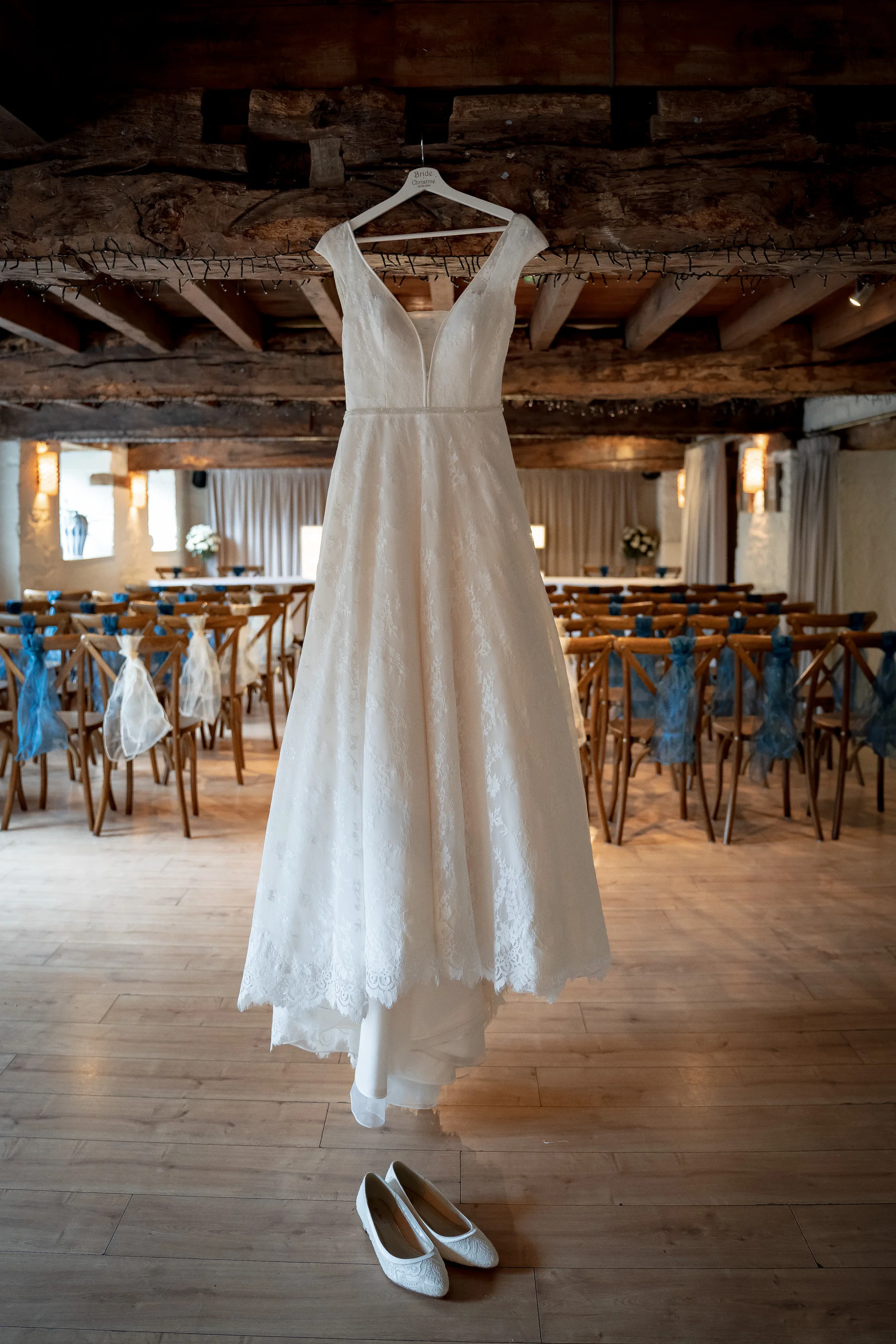 An ivory lace a line wedding dress hangs from a wooden beam inside a rustic barn ceremony venue, with matching bridal flats placed neatly on the wooden floor below.
