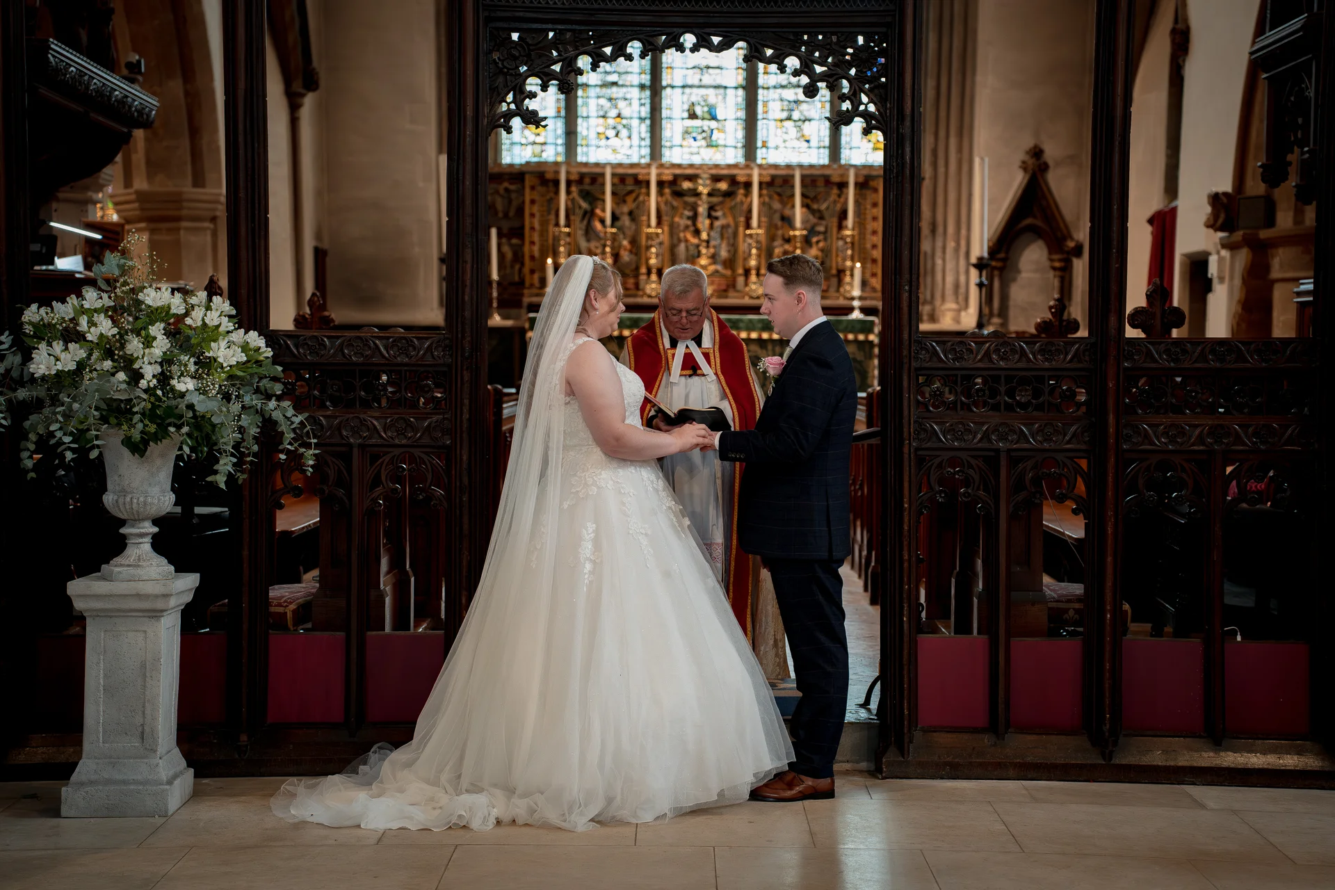 The bride and groom exchange rings during their traditional church ceremony, framed by ornate wooden rood screen carvings and stained glass windows.