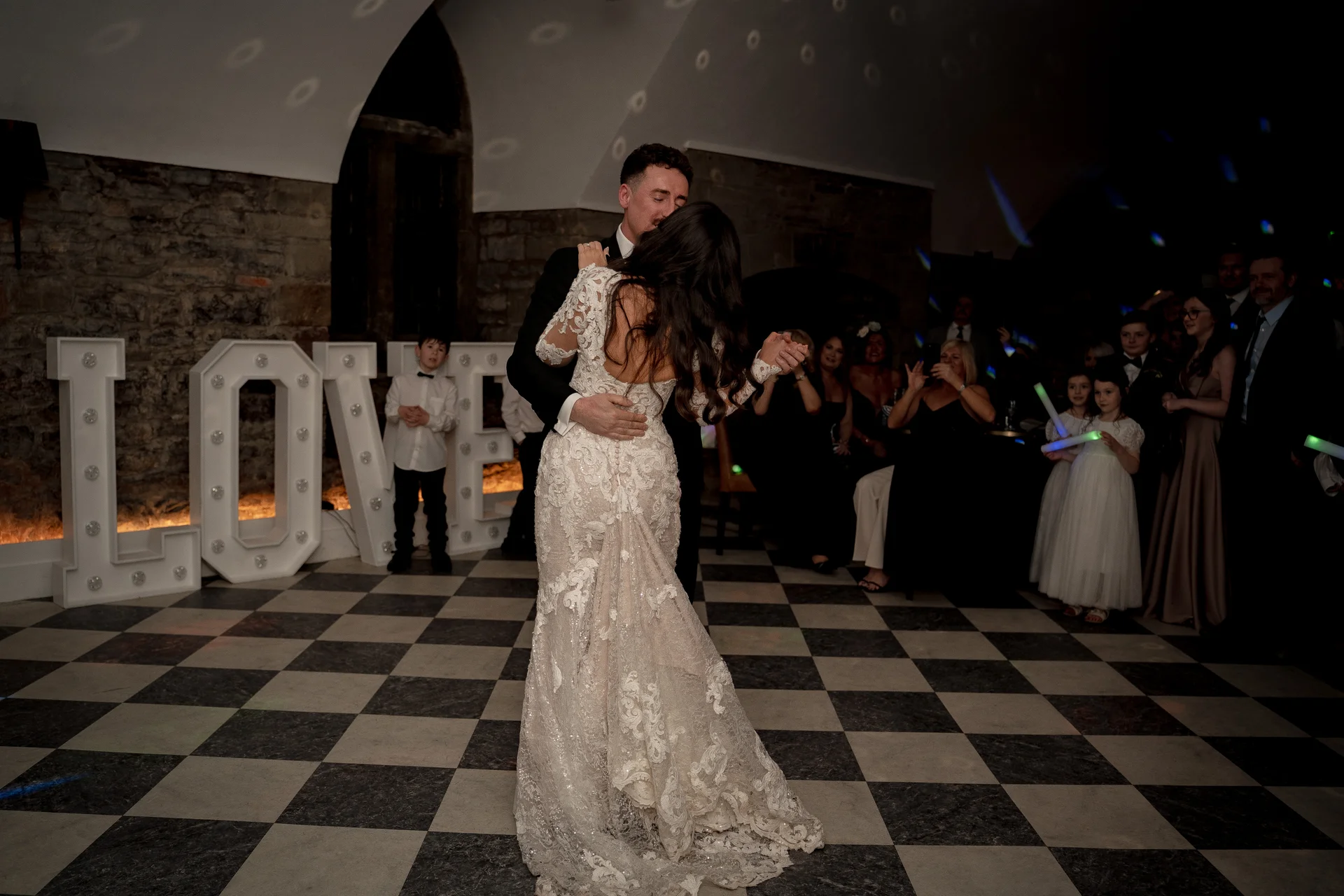 A newlywed couple shares a tender kiss during their first dance on a black and white checkered floor in a stone barn venue.