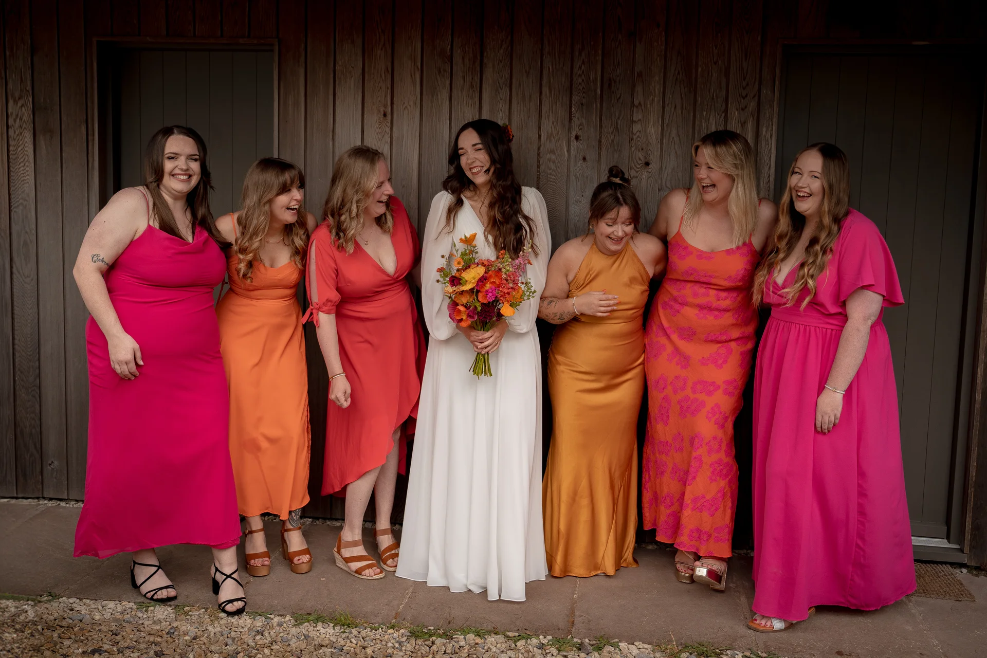 A bride in a white long-sleeved gown holds a vibrant bouquet of orange and pink flowers, laughing alongside her bridesmaids who wear mismatched colorful dresses against a wooden barn wall.