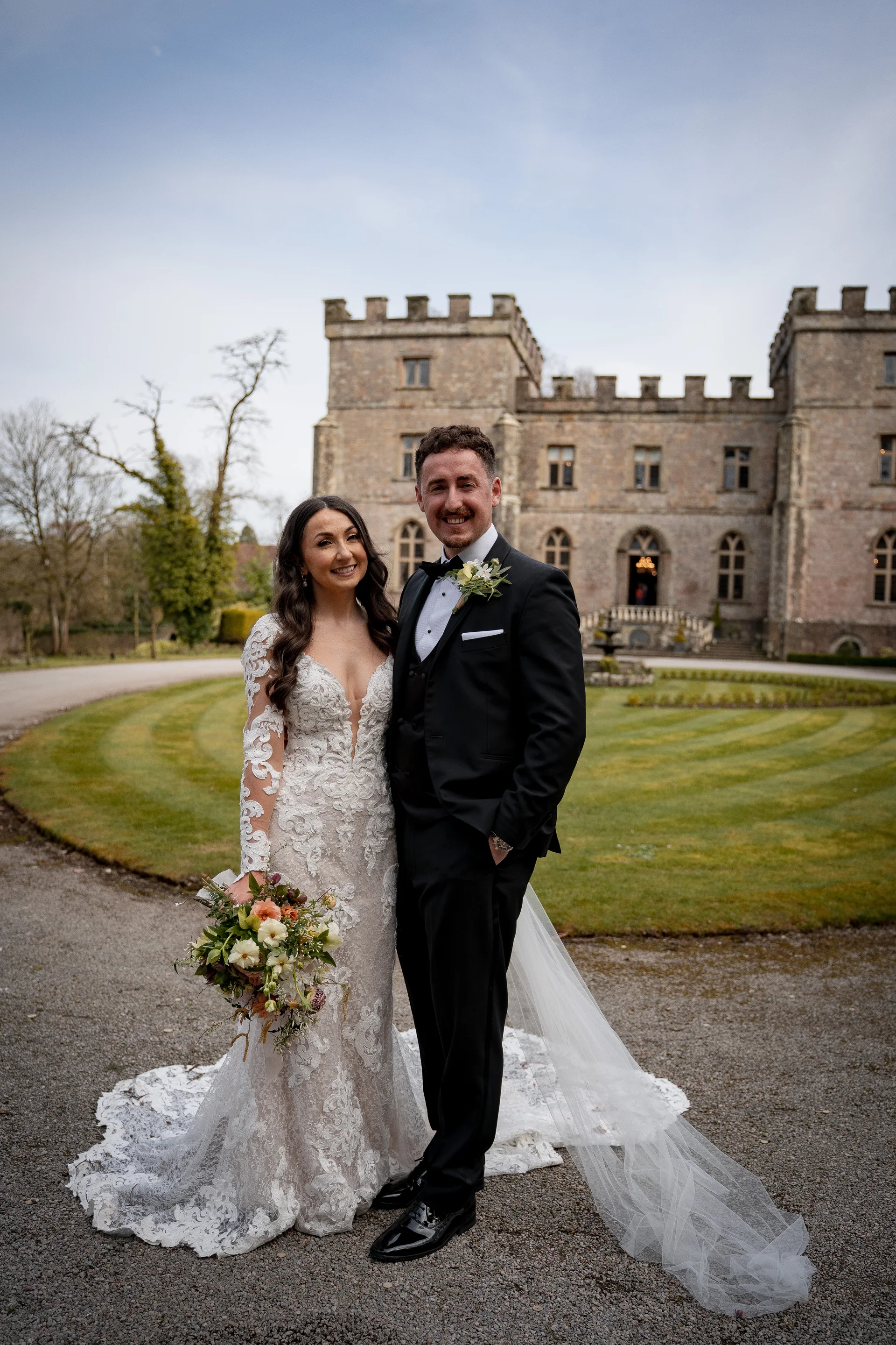 A newlywed couple poses smiling in front of a majestic stone castle with manicured lawns on their wedding day.