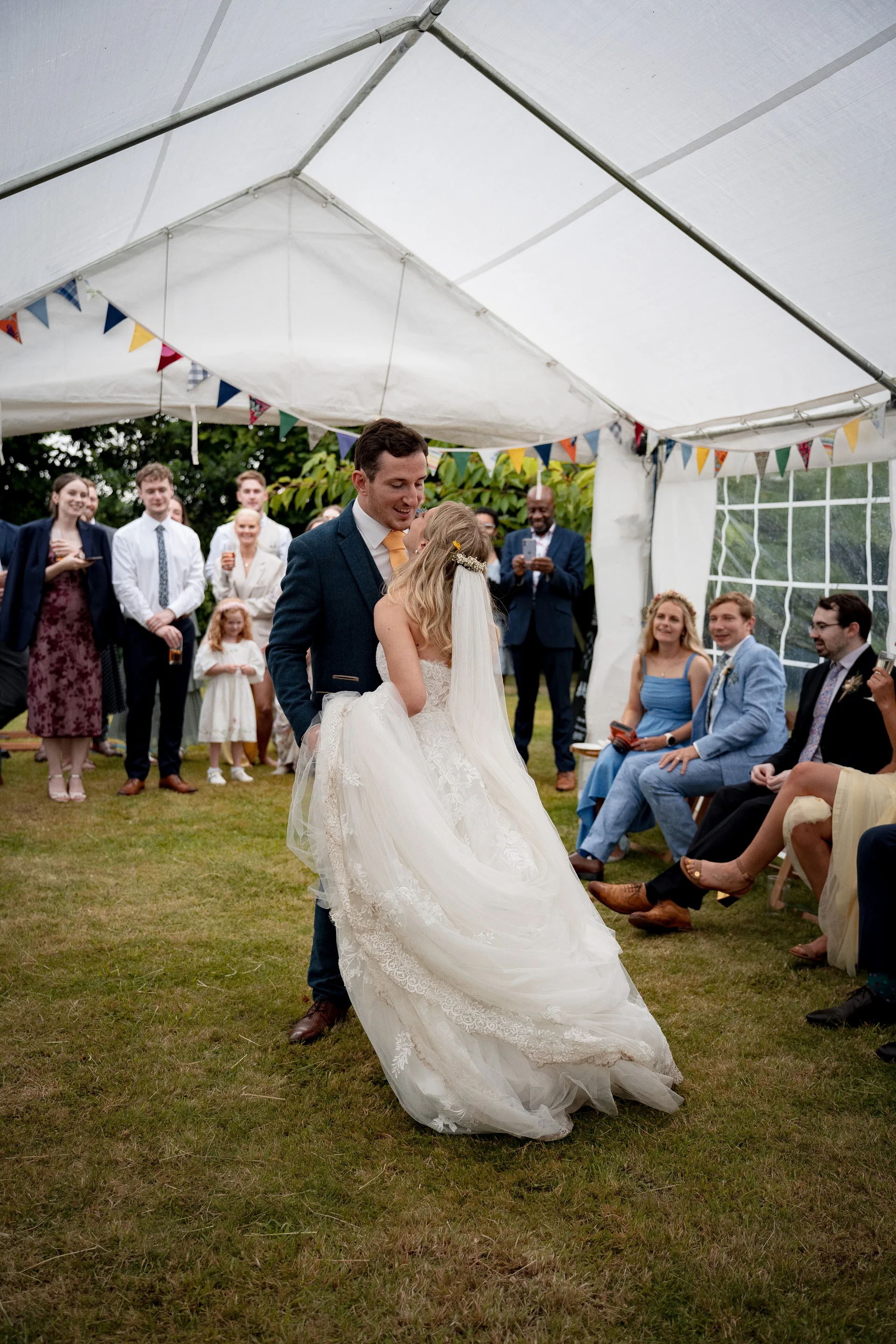 A newlywed couple share their first dance under a white marquee adorned with colourful bunting, surrounded by seated and standing guests on a grassy lawn.